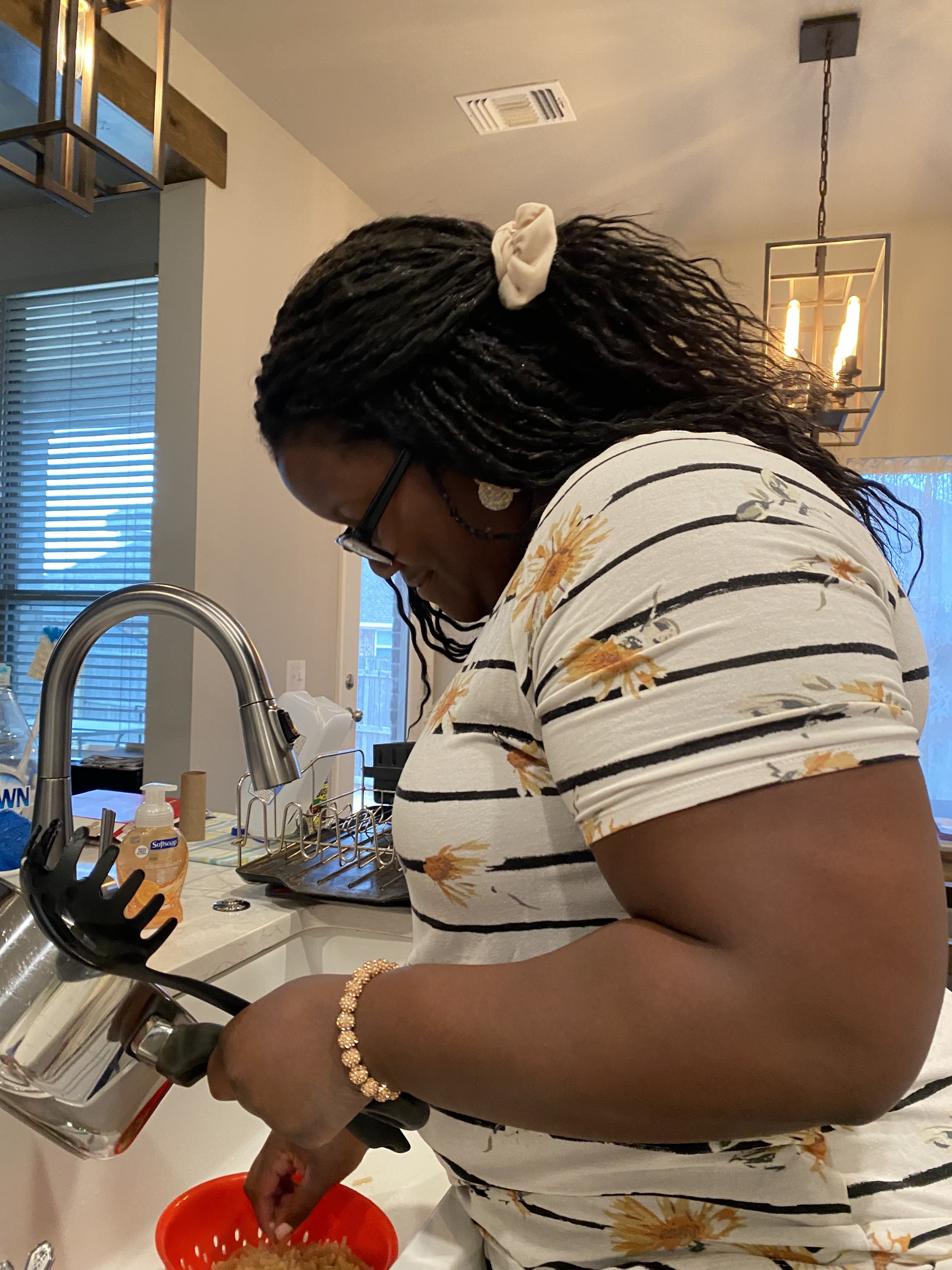 A woman with dark hair styled in twists, wearing glasses, a floral and striped shirt, a gold bracelet, and heart-shaped earrings, is standing at a kitchen sink, pouring rice from a silver rice cooker into a red colander. The kitchen has a faucet, soap dispenser, and various items on the countertop, with a window with blinds and a hanging light fixture in the background.
