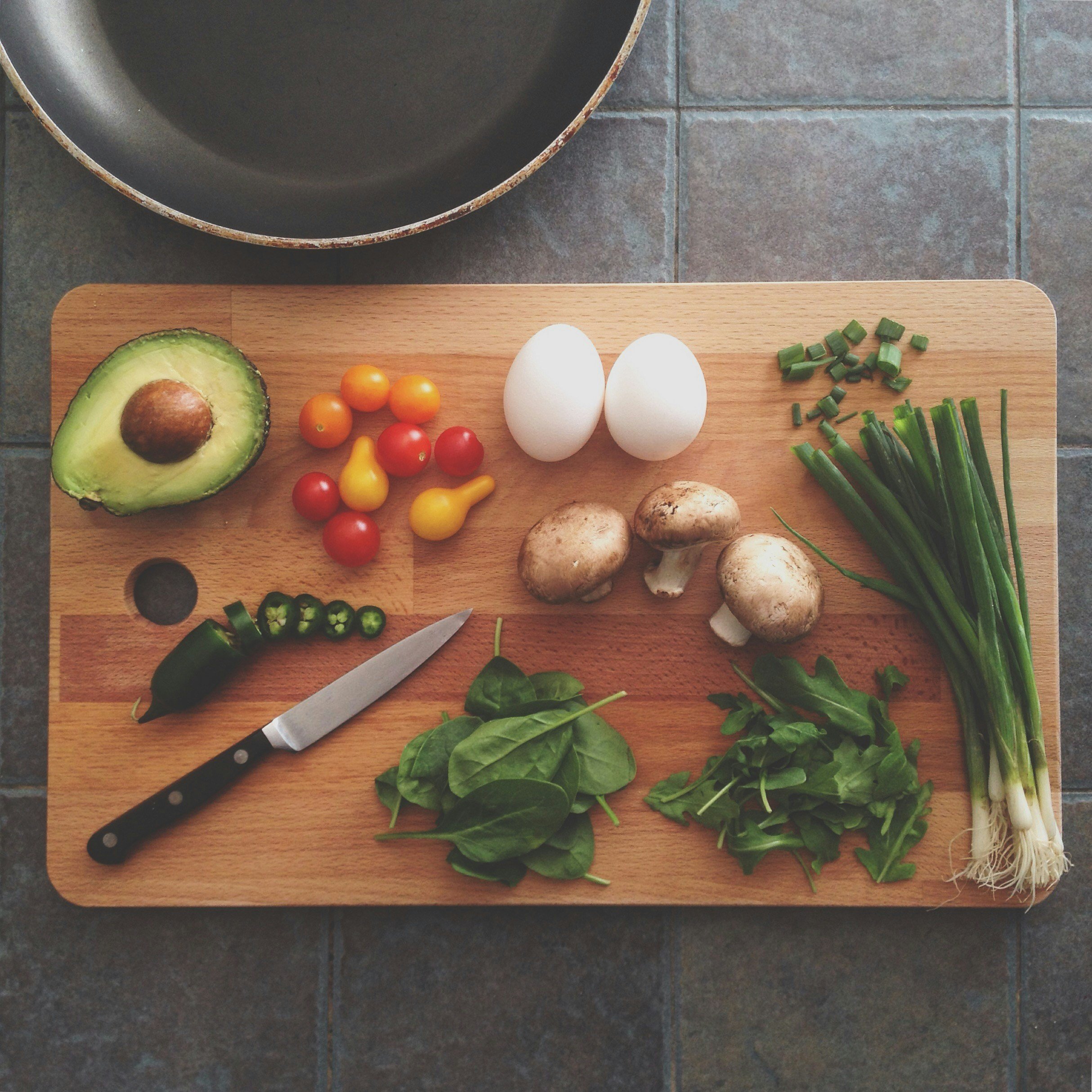 A wooden cutting board with various fresh ingredients, including an avocado, cherry tomatoes, eggs, mushrooms, chopped green onions, sliced jalapeño, spinach, and arugula, alongside a knife, on a gray tiled surface.