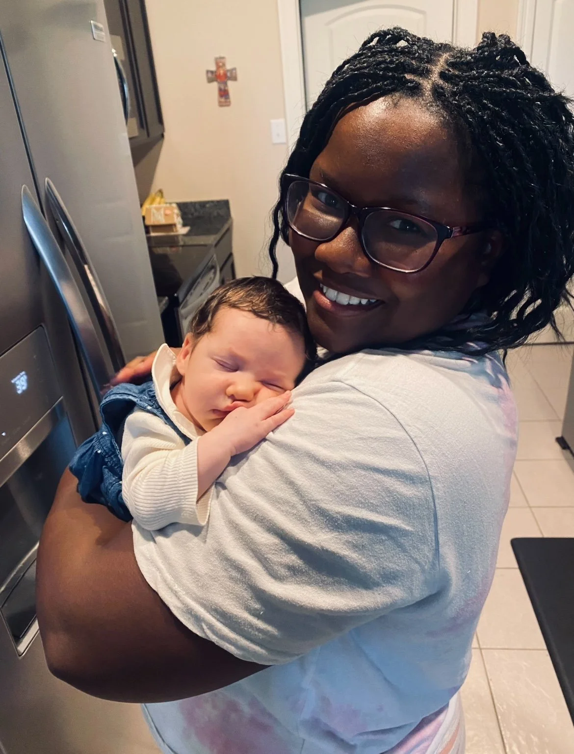A woman with glasses holding a sleeping baby in a kitchen.