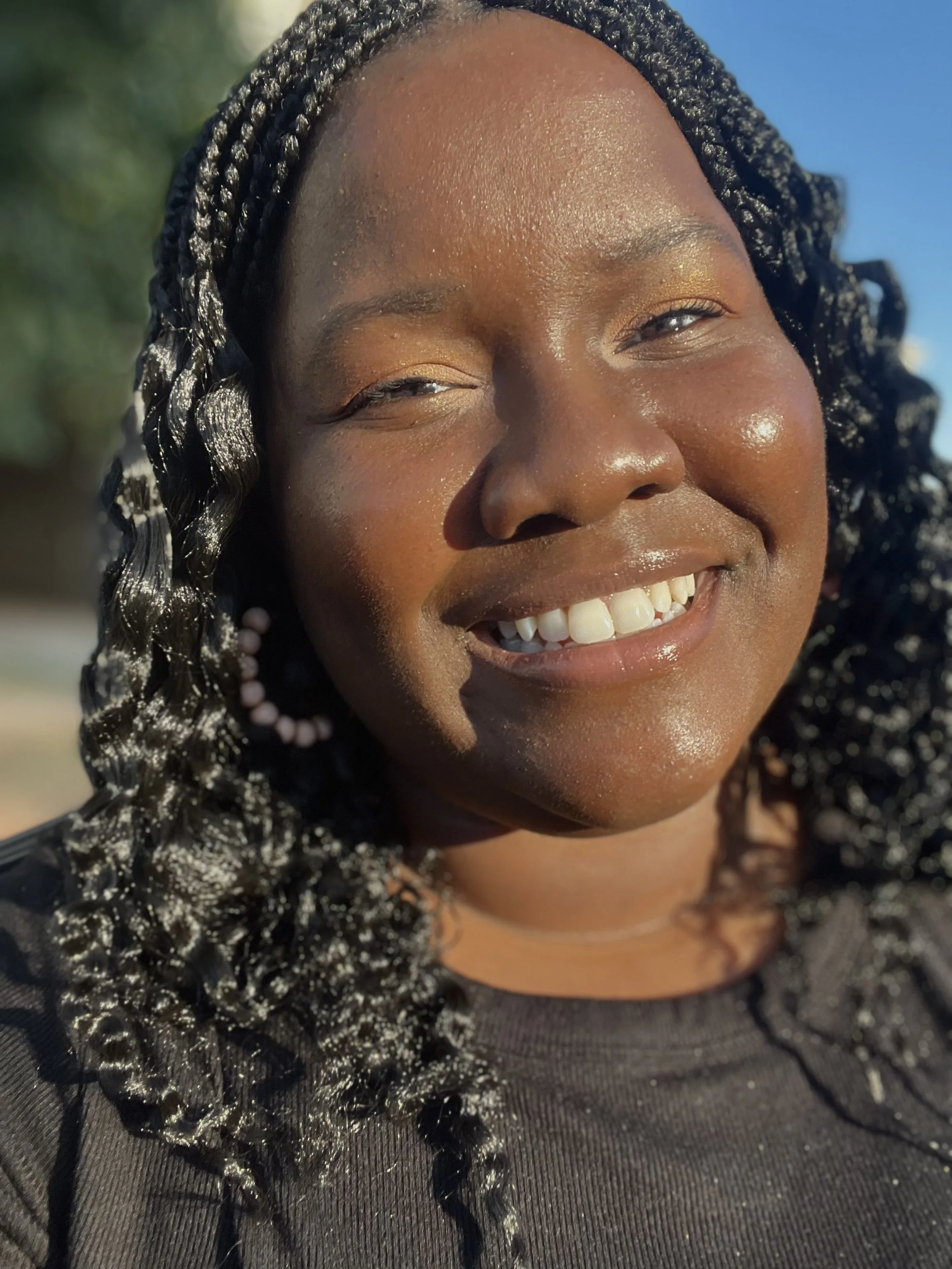 A woman smiling outdoors with sunlight shining on her face, curly hair, wearing earrings and a black top.