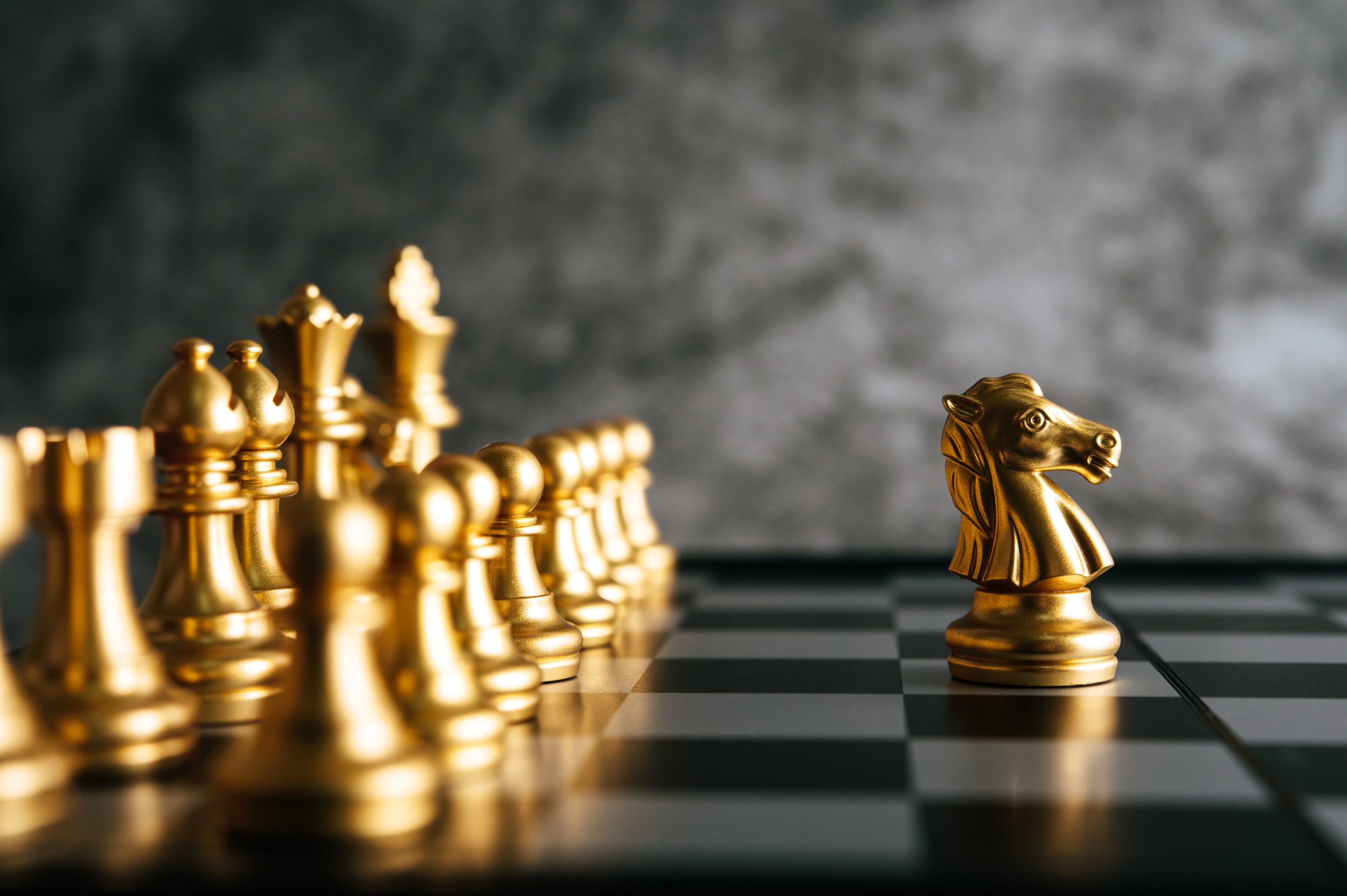 Gold chess pieces, with a knight in focus on a black and white chessboard, facing an array of other pieces in the background, against a textured gray backdrop.
