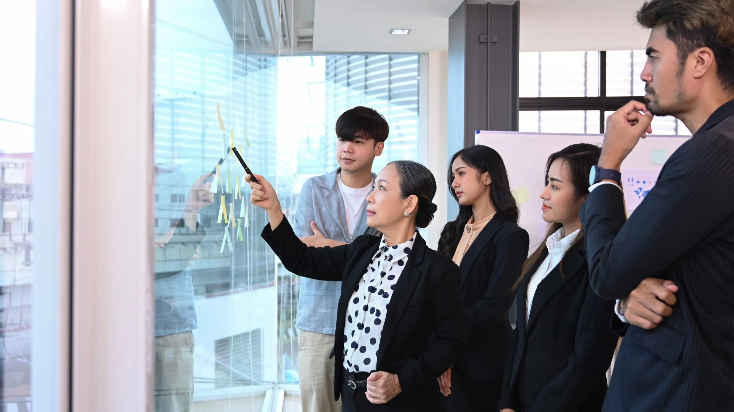 A group of diverse professionals, including women and men, are having a discussion in an office with large glass windows. One woman is pointing at sticky notes on the glass wall, while others watch attentively.