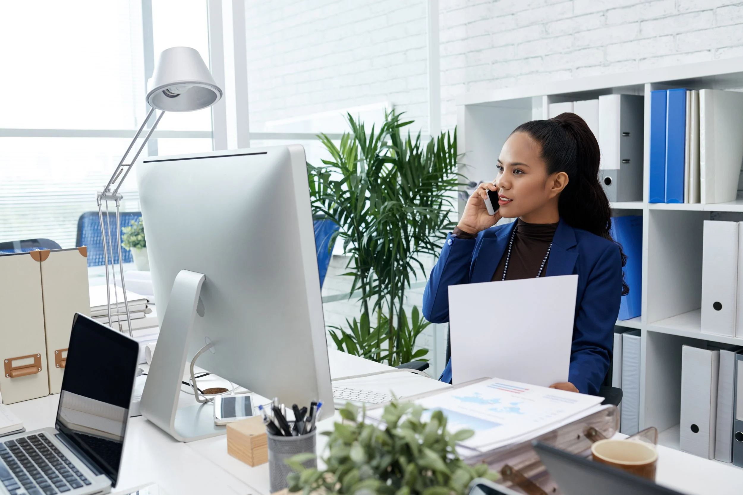 woman in blue blazer talking on phone at her office desk, surrounded by computer, laptop, folders, and plants.