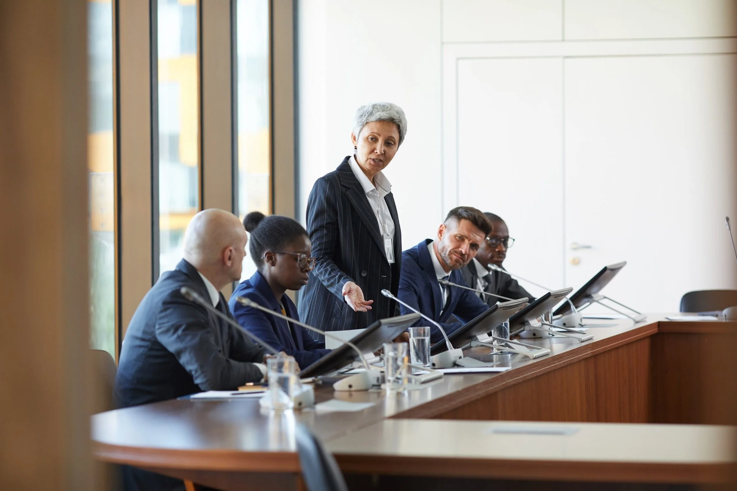 A diverse group of professionals seated at a conference table, with a woman standing and speaking during a meeting in a modern office setting.