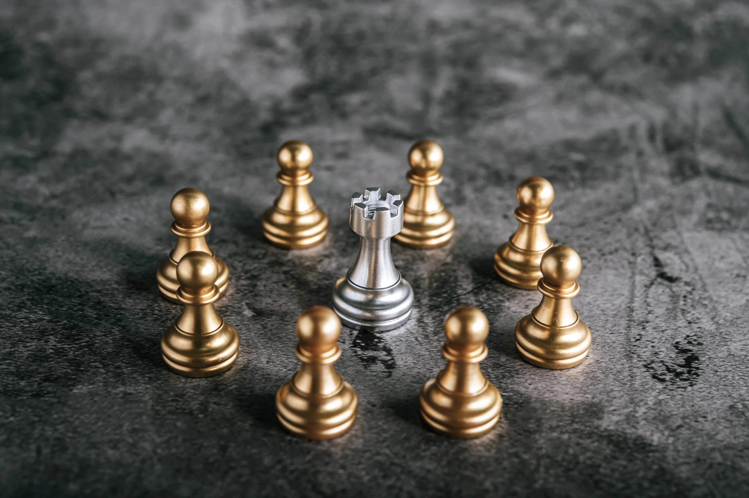 A silver king chess piece surrounded by nine gold pawns on a dark textured surface.