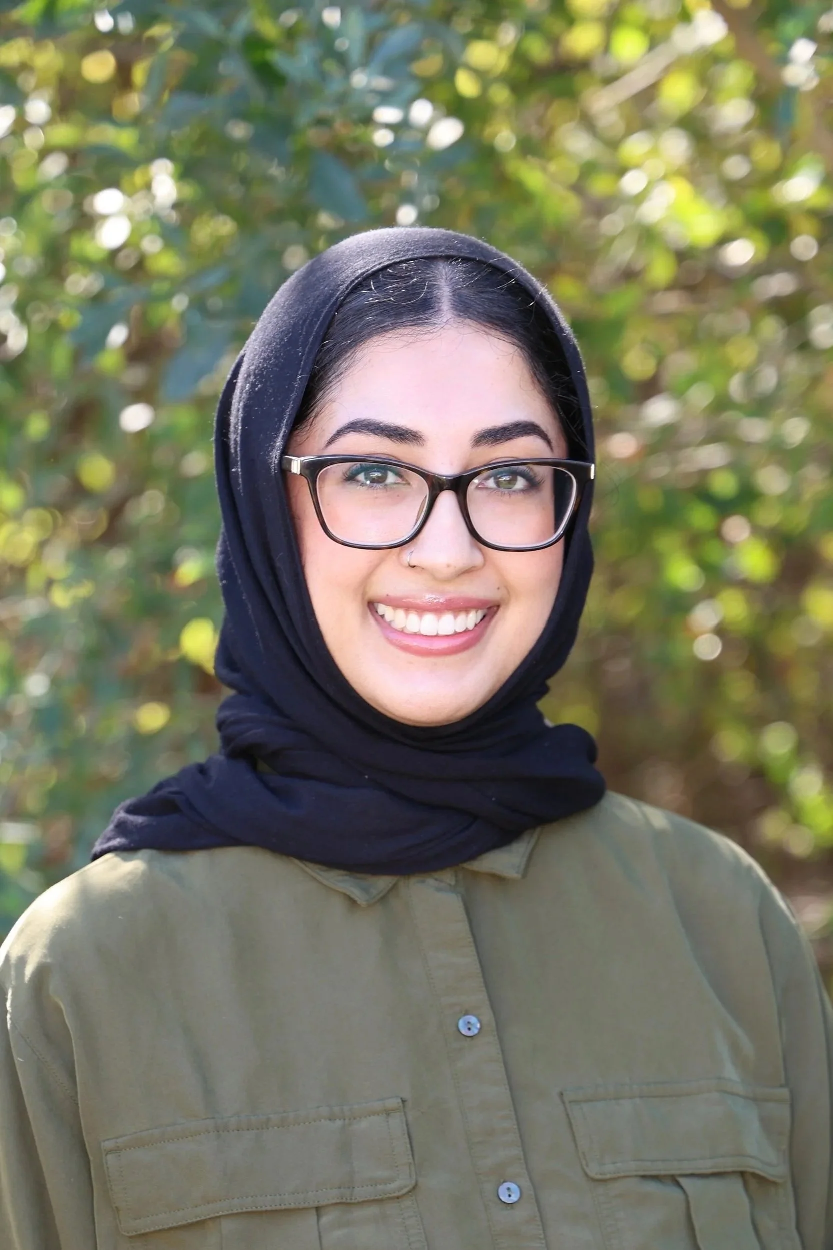 A headshot of a smiling woman wearing a black hijab, black glasses, and a khaki green shirt