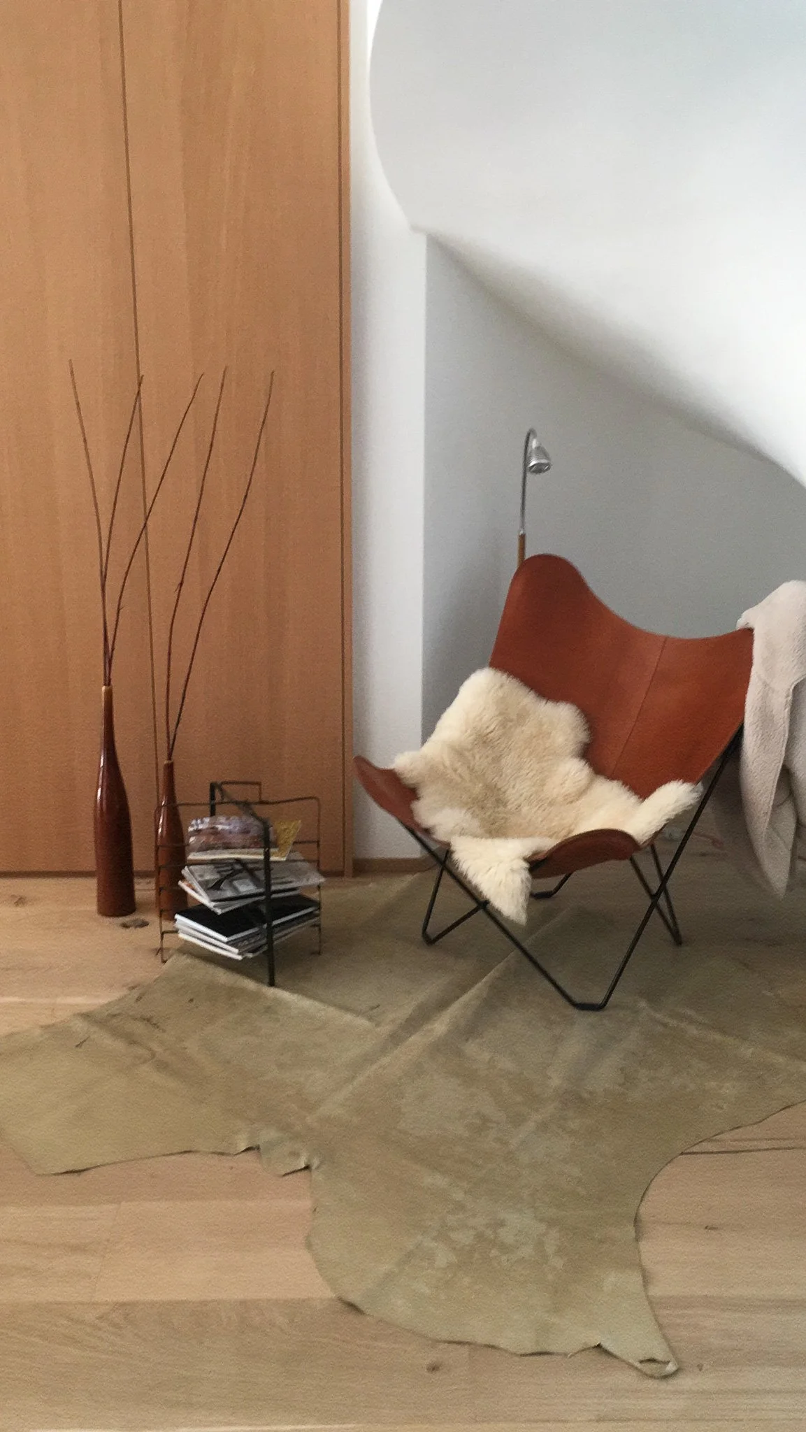 Un coin de salon avec une chaise en cuir brun, un tapis beige en peau de vache, une étagère avec des magazines, deux grands vases en terre cuite, une lampe de lecture, et une couverture beige sur le fauteuil.