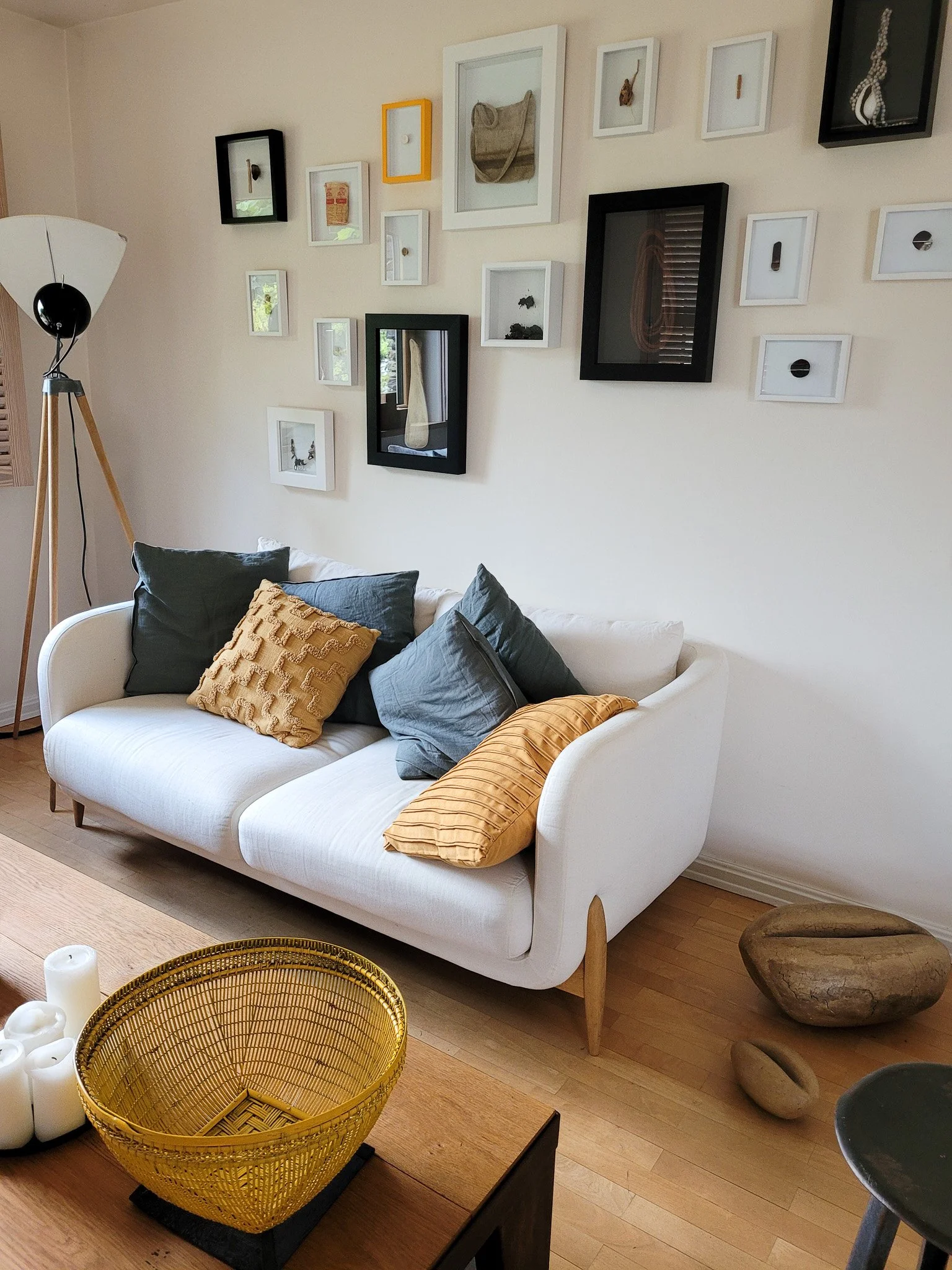 Un salon avec un canapé blanc et plusieurs oreillers gris et jaunes, un mur décoré de plusieurs cadres photo de différentes tailles, une lampe sur pied en bois à gauche, une table en bois avec un panier jaune et plusieurs bougies, et des pierres décoratives sur le sol à droite.
