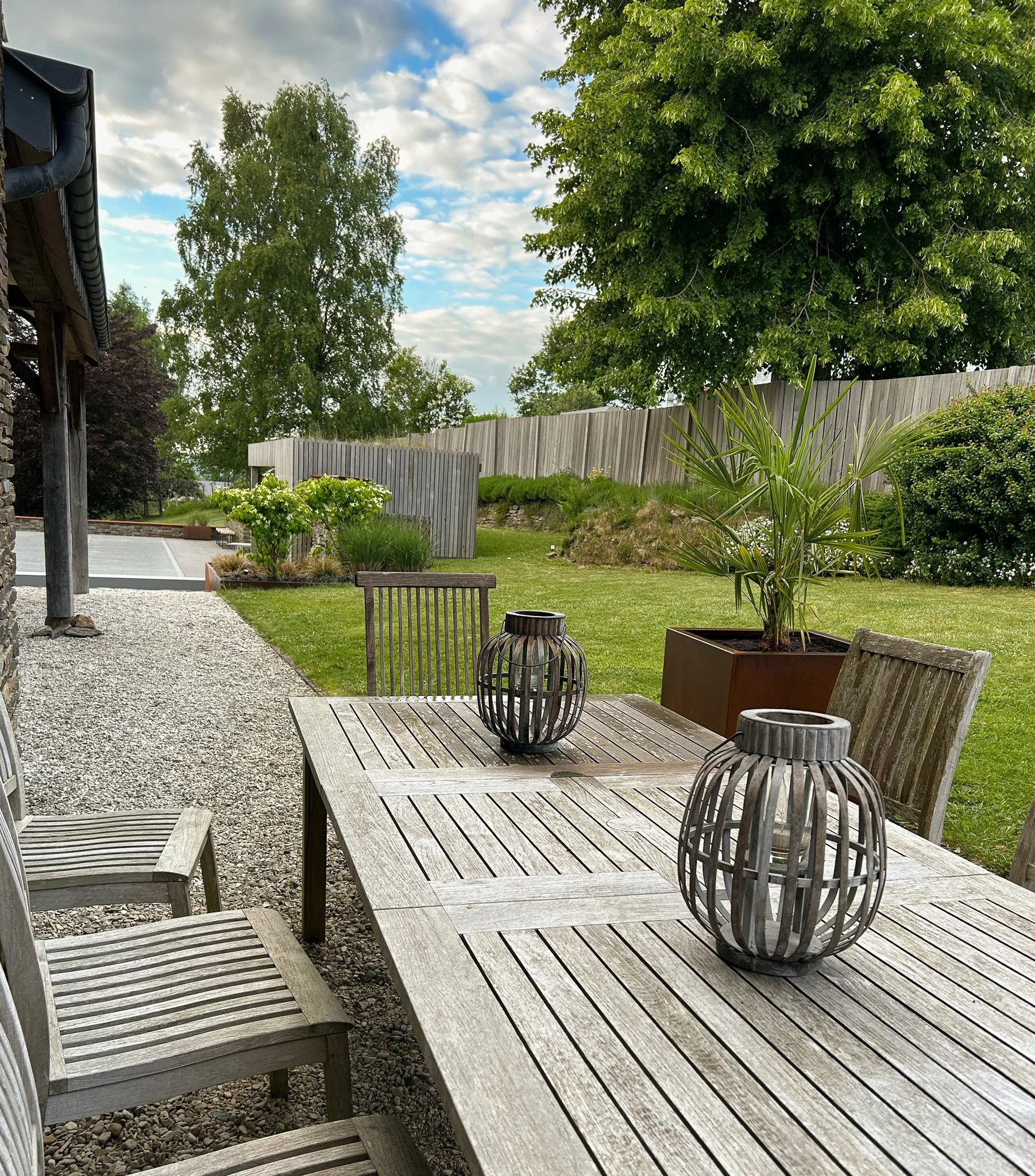 Une terrasse extérieure en pierre et en bois avec une table en bois, entourée de chaises en bois, décorée de deux lanternes en bois, avec des plantes en pot vertes et un jardin bien entretenu avec des arbres en arrière-plan, sous un ciel partiellemen