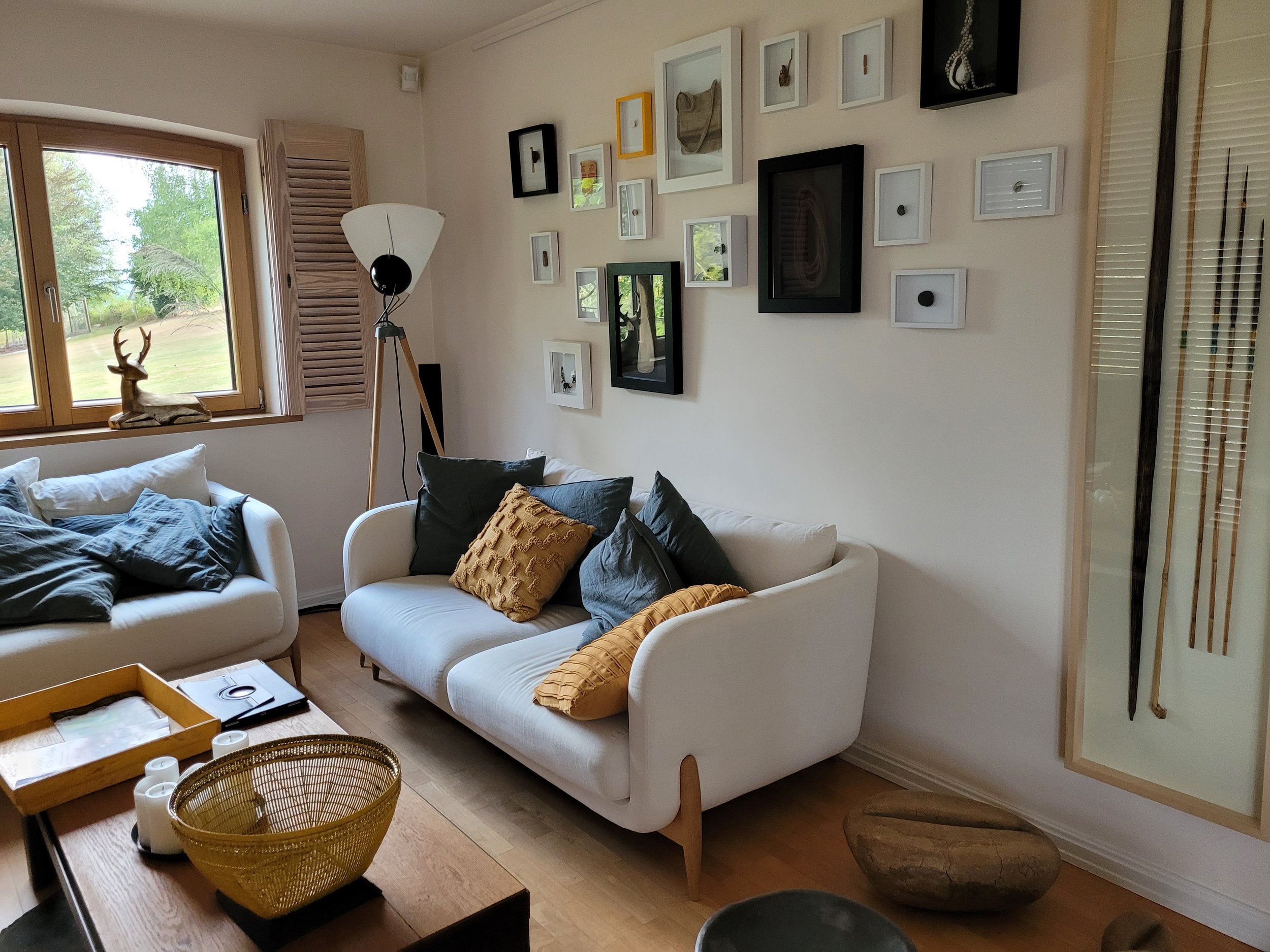 Salon avec deux canapés blancs, plusieurs coussins, une table en bois avec un panier en osier, une lampe sur pied, plusieurs cadres décoratifs sur le mur, une fenêtre montrant un extérieur vert, des objets décoratifs dans un cadre en verre sur le mur