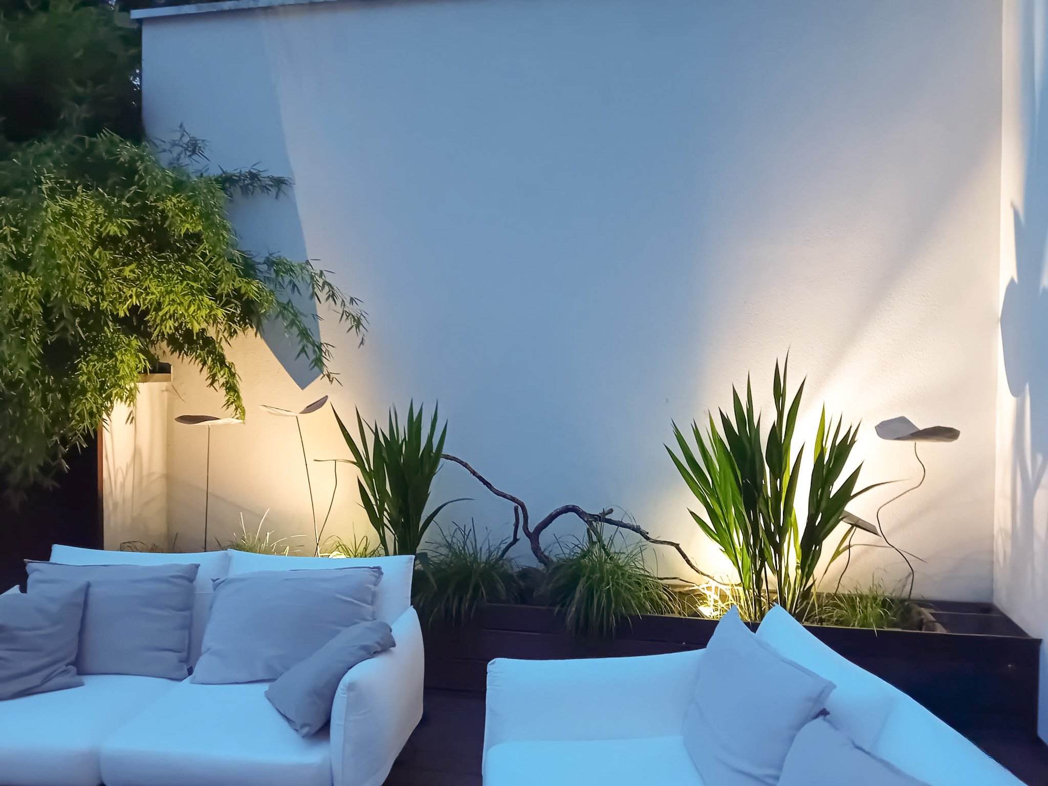 Terrasse avec deux canapés blancs et des coussins gris, plantes vertes et éclairage d'ambiance