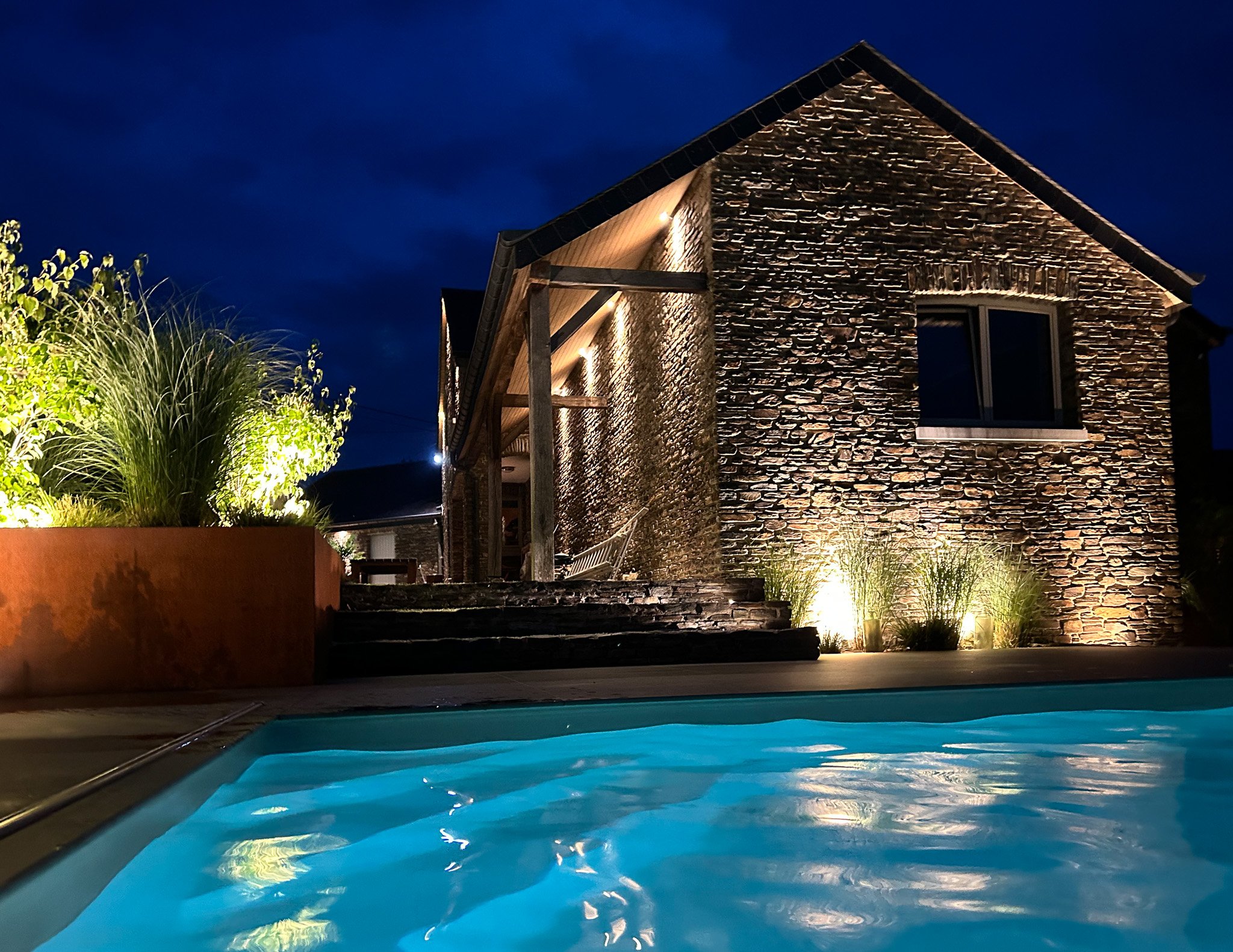 Une maison en pierre éclairée la nuit, avec une piscine en avant-plan et des plantes lumineuses autour.