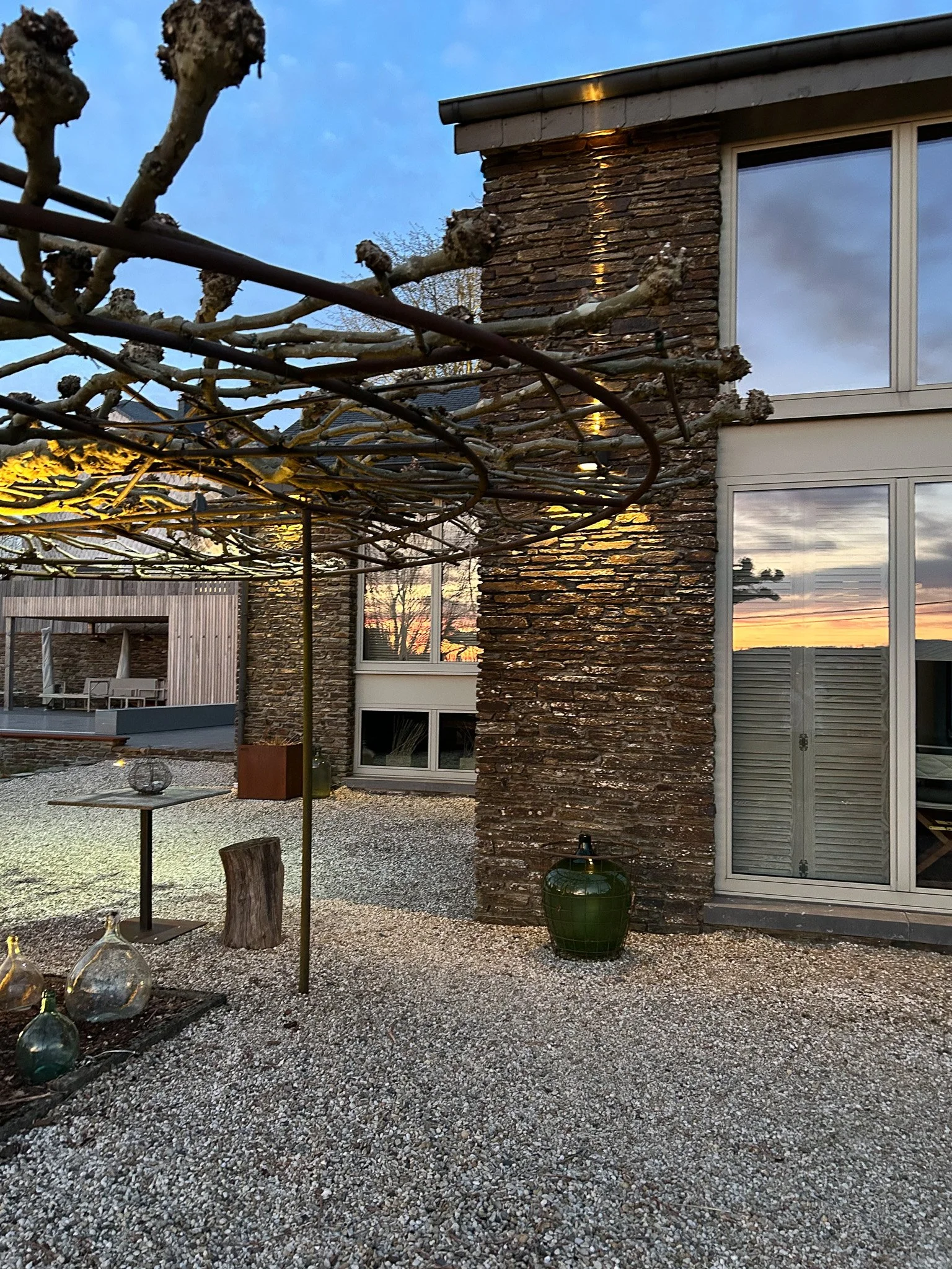 vue d'une terrasse extérieure avec un sol en gravier, une maison moderne en pierre et en verre, et des arbres taillés sans feuilles, lors du coucher de soleil.