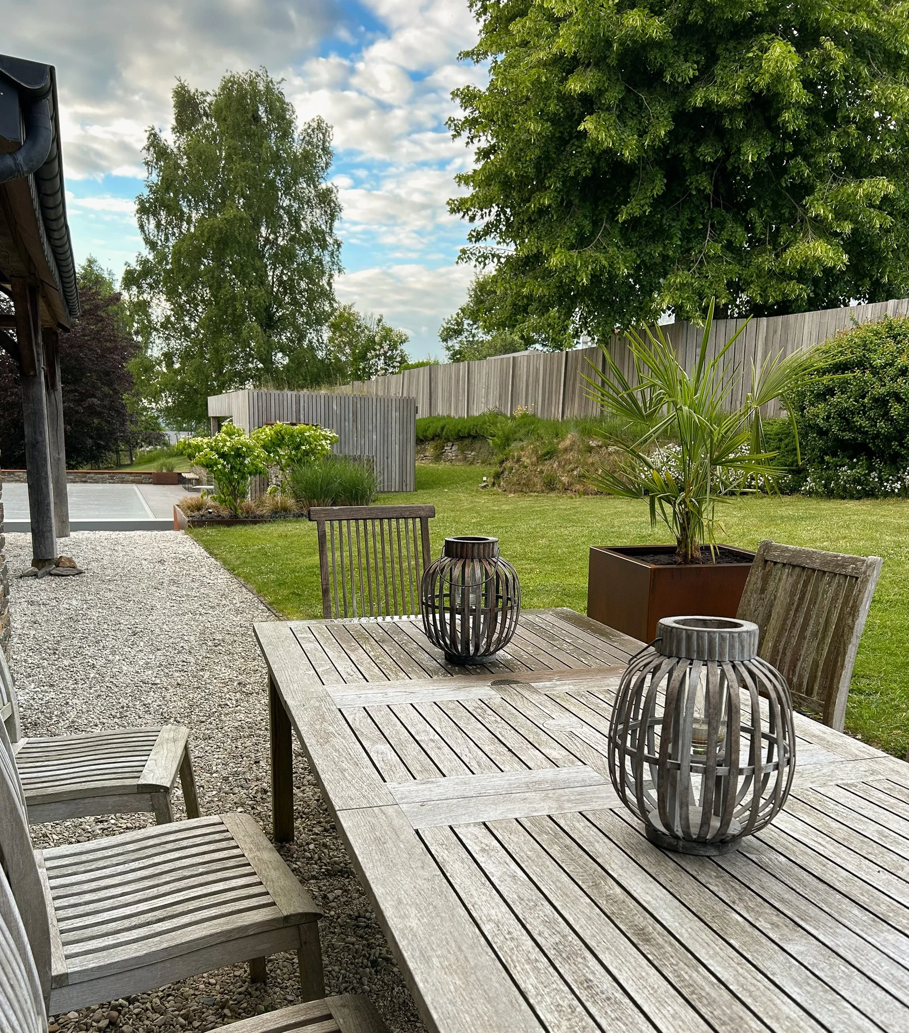 Une terrasse en bois avec des chaises en bois, une table en bois, et deux lanternes décoratives en métal. En arrière-plan, un jardin avec des arbres, une pelouse verte, et un ciel partiellement nuageux.