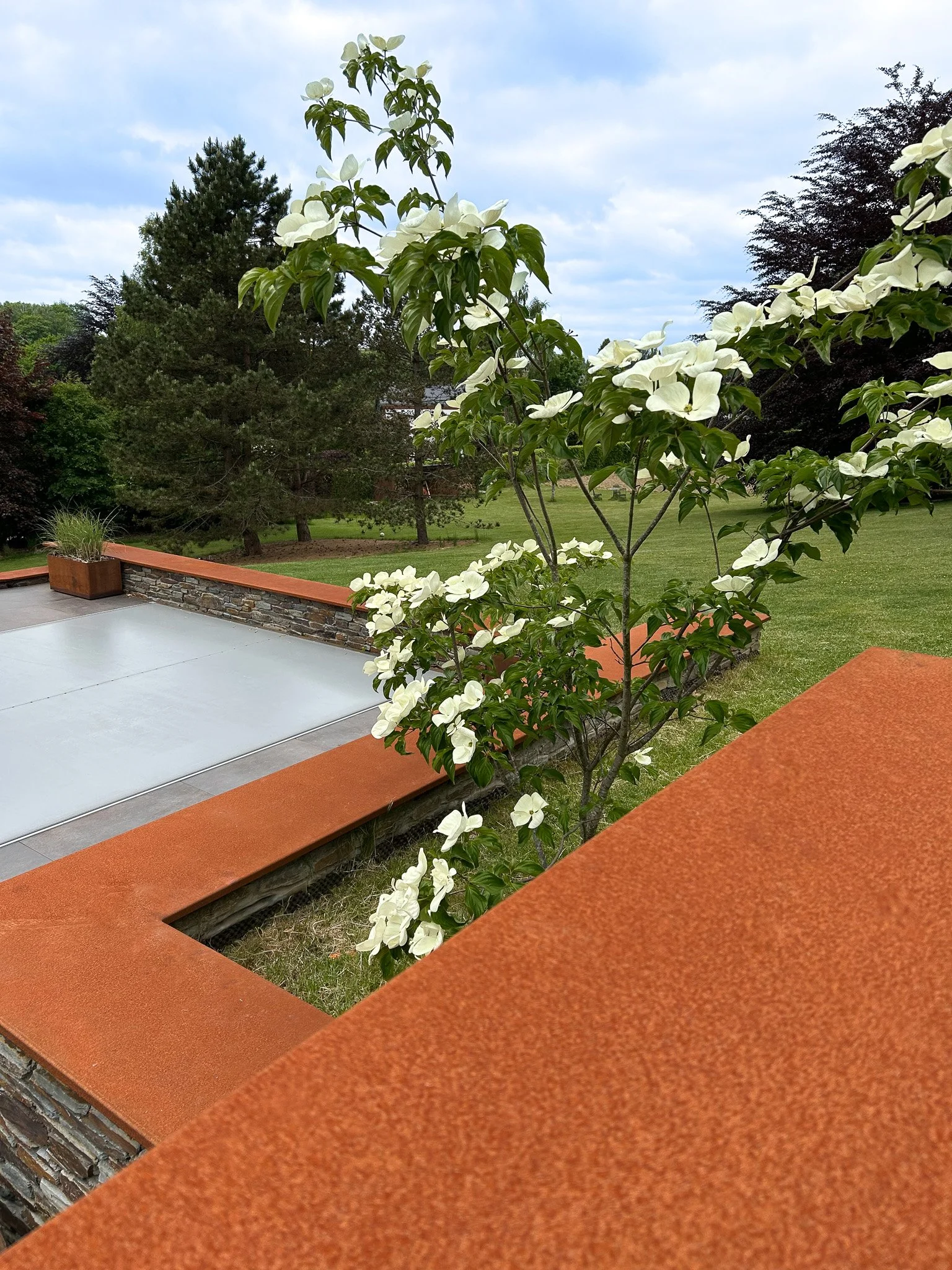 Vue d'une terrasse avec une balustrade en pierre, une table ou un plancher en béton, et un arbre en fleurs blanches dans un jardin verdoyant avec des arbres et le ciel nuageux en arrière-plan.