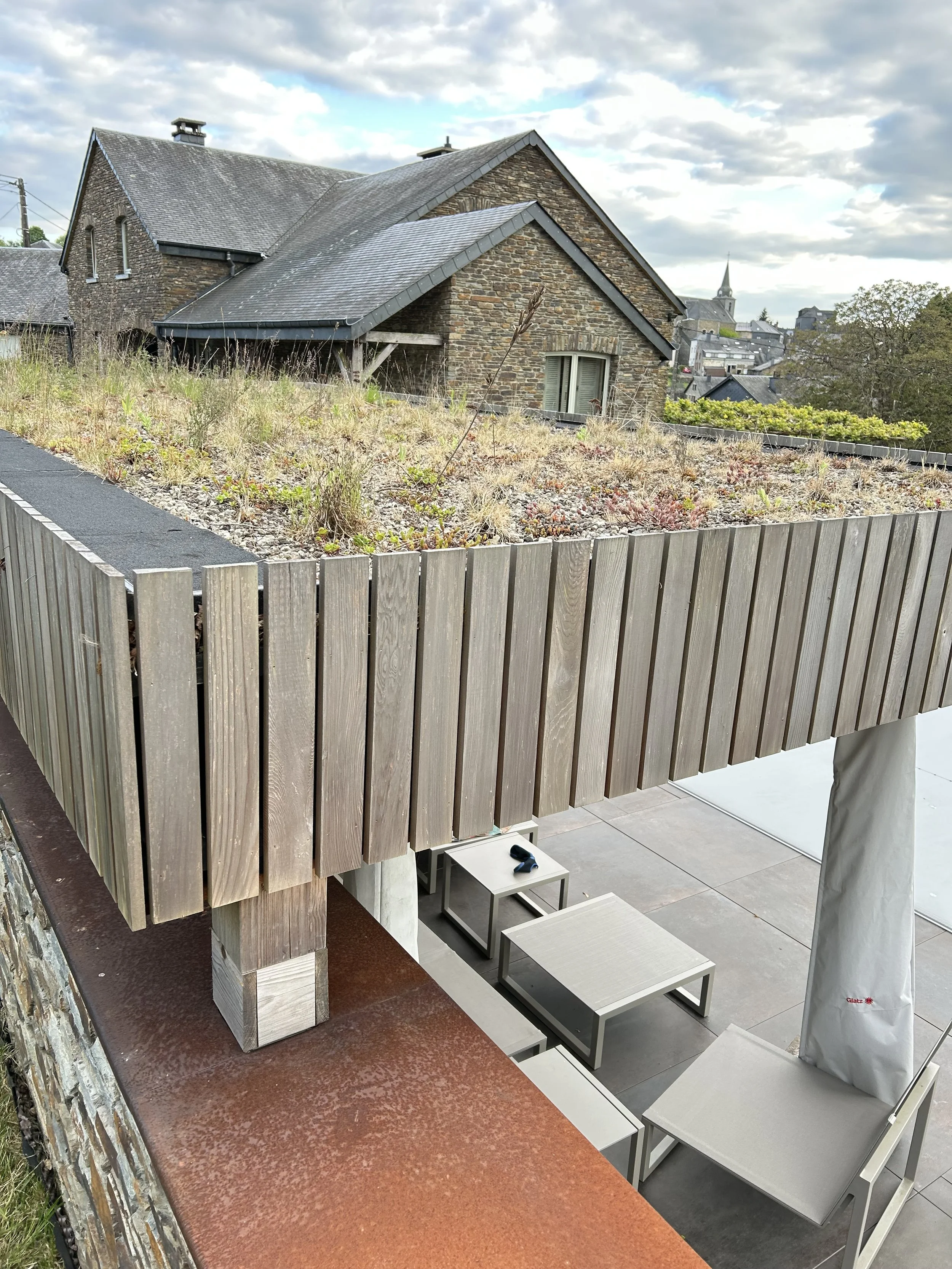 Photo d'une terrasse avec mobilier en métal gris clair, comprenant une table et des fauteuils, en dessous d'une clôture en bois clair. Au-delà, on voit un jardin sans gazon, avec des plantes vivaces, et en arrière-plan une maison en pierre avec un to