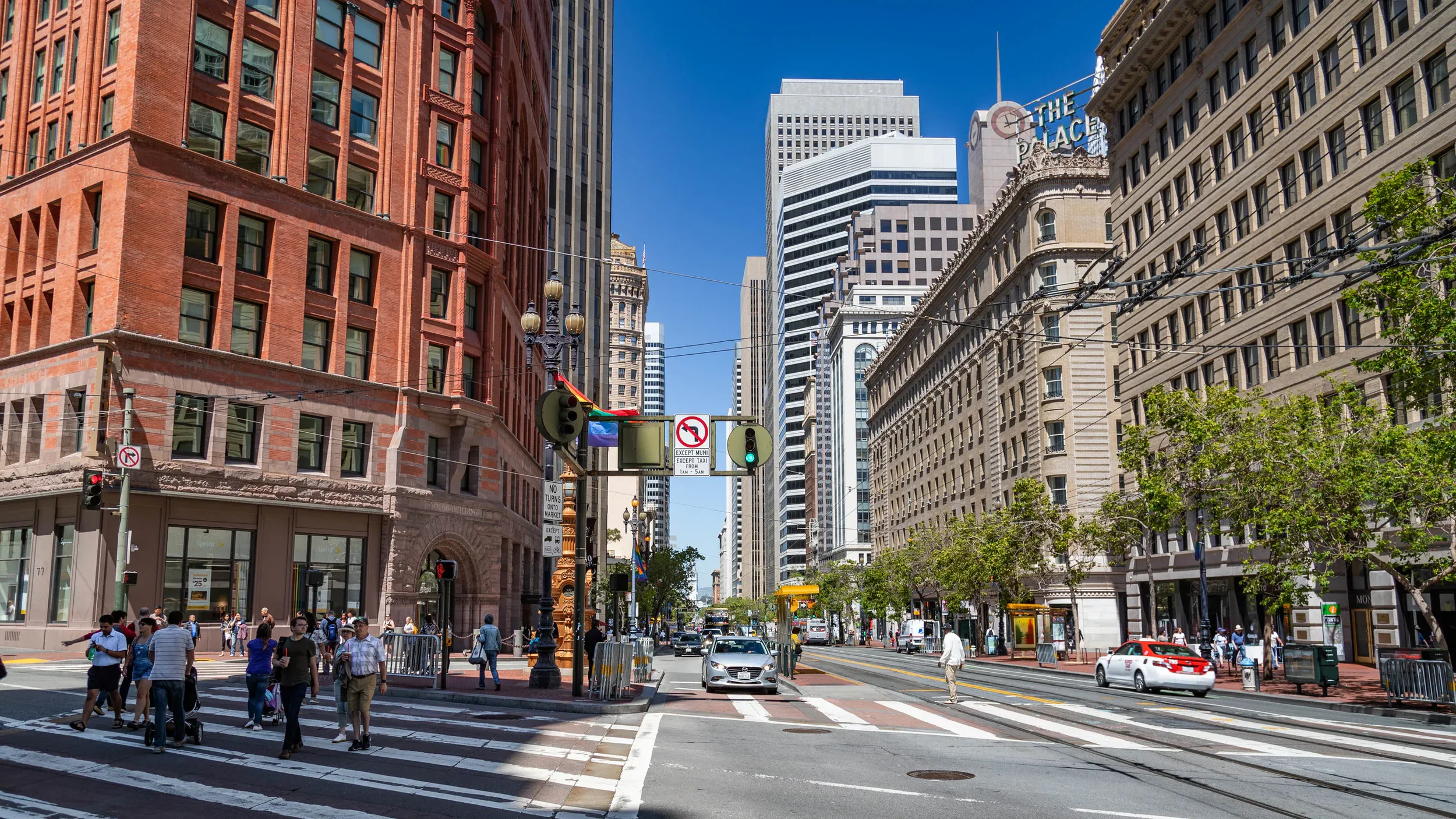 A busy city street with pedestrians crossing at a crosswalk, several cars on the road, tall buildings on both sides, and clear blue sky overhead.