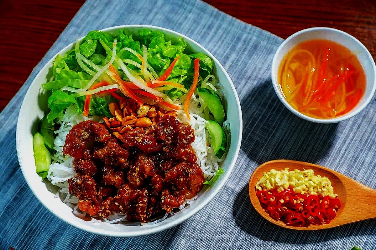 A bowl of salad with greens, shredded carrots, and peanuts, topped with a meat sauce. Accompanying are a small cup of do chua pickled vegetables and a wooden spoon with chopped garlic and red chili peppers.