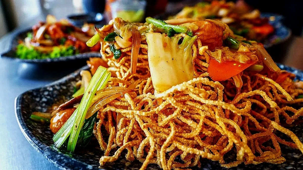 Close-up of a plate of crispy fried noodles with vegetables, including cabbage, carrots, and green onions, served on a black textured plate.
