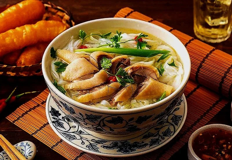 A bowl of Vietnamese pho with slices of meat, herbs, and green onion, on a wooden placemat with side dishes and a drink in the background.