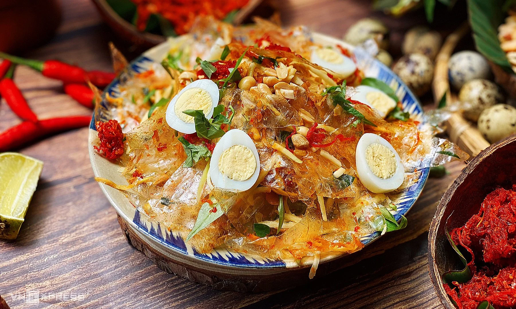 A bowl of Vietnamese shrimp and pork jello salad garnished with sliced boiled eggs, scallions, chopped peanuts, and red chili flakes, placed on a rustic wooden table with side dishes and chili peppers around it.
