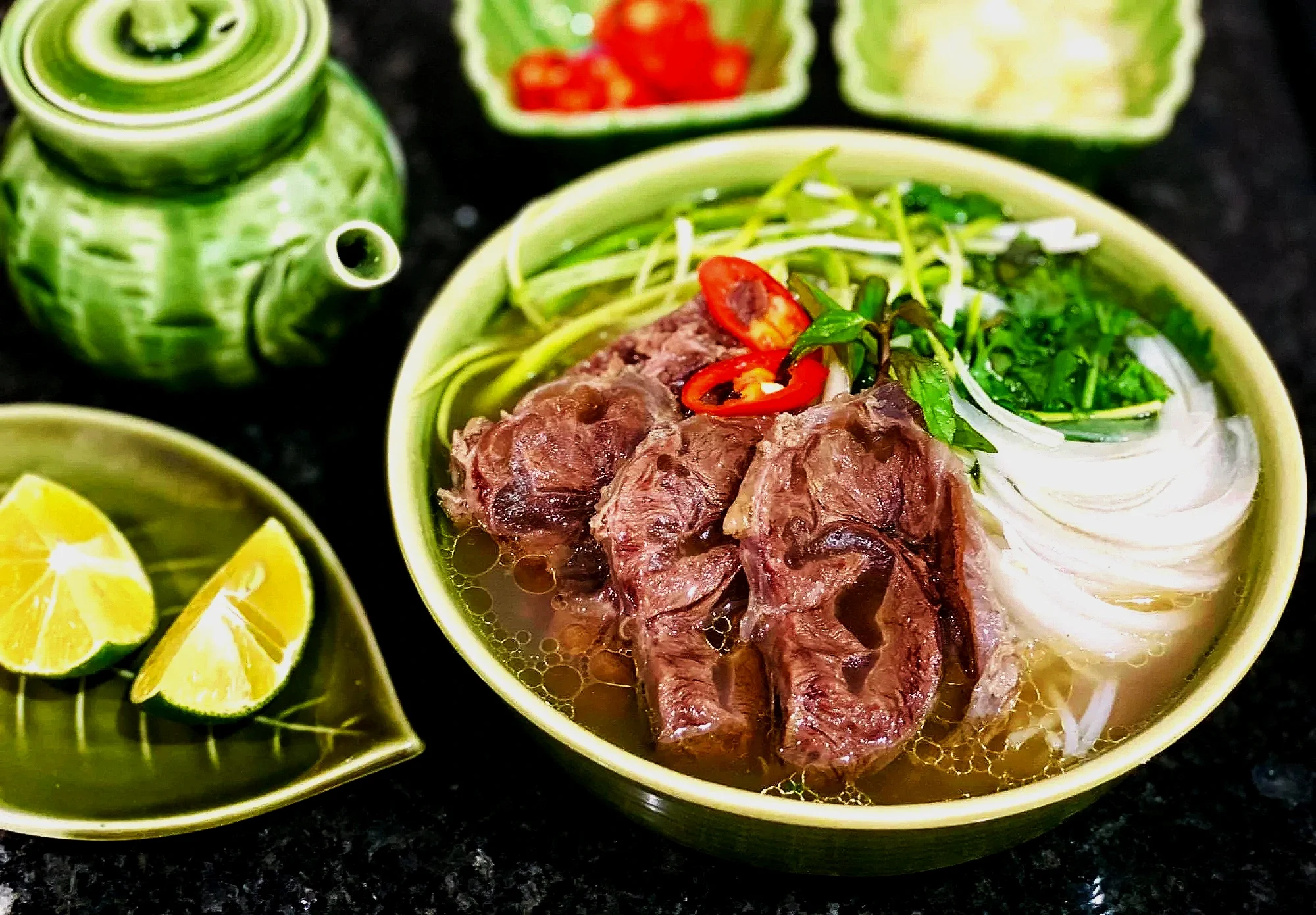 Vietnamese pho soup with beef, rice noodles, and herbs in a bowl, accompanied by lime slices and green leafy vegetables, on a black countertop.