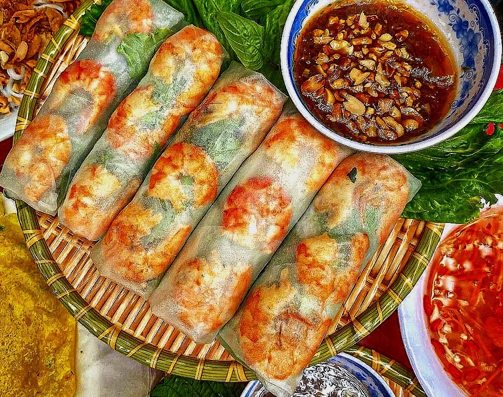 Five Vietnamese spring rolls served on a basket with fresh herbs, accompanied by a bowl of dipping sauce with chopped nuts.