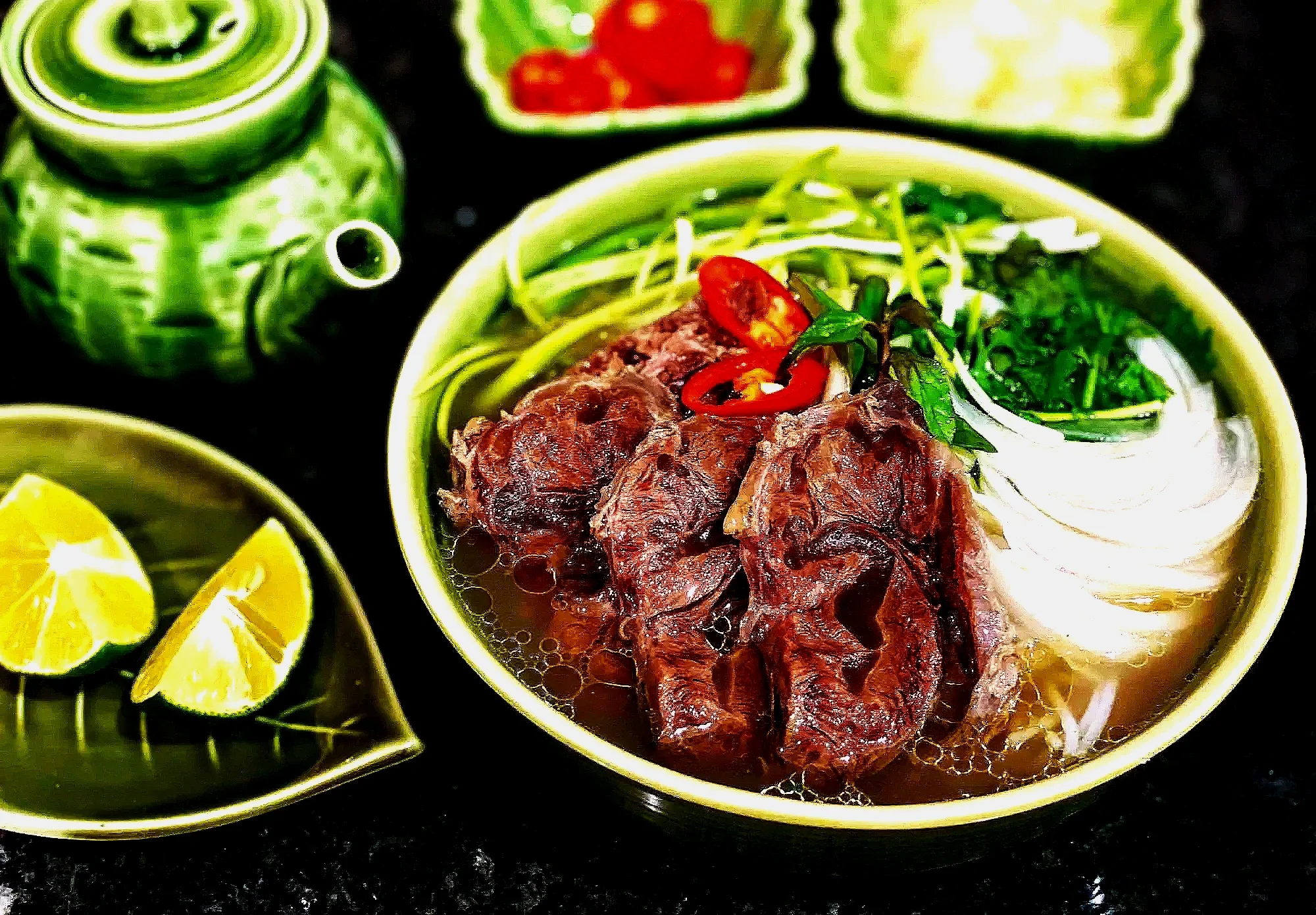 A bowl of Vietnamese pho featuring beef, rice noodles, sliced chili peppers, and green herbs, with lemon wedges, a teapot, and condiment containers in the background.