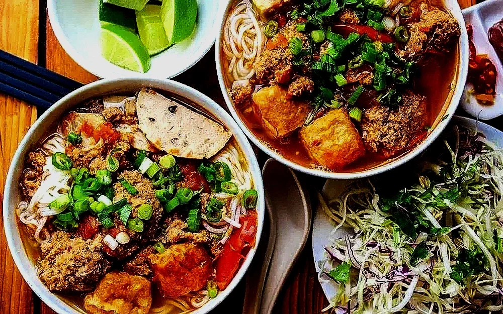 Two bowls of Vietnamese noodle soup with meat, green onions, and spices, served with lime wedges, and a side of shredded cabbage salad with herbs.