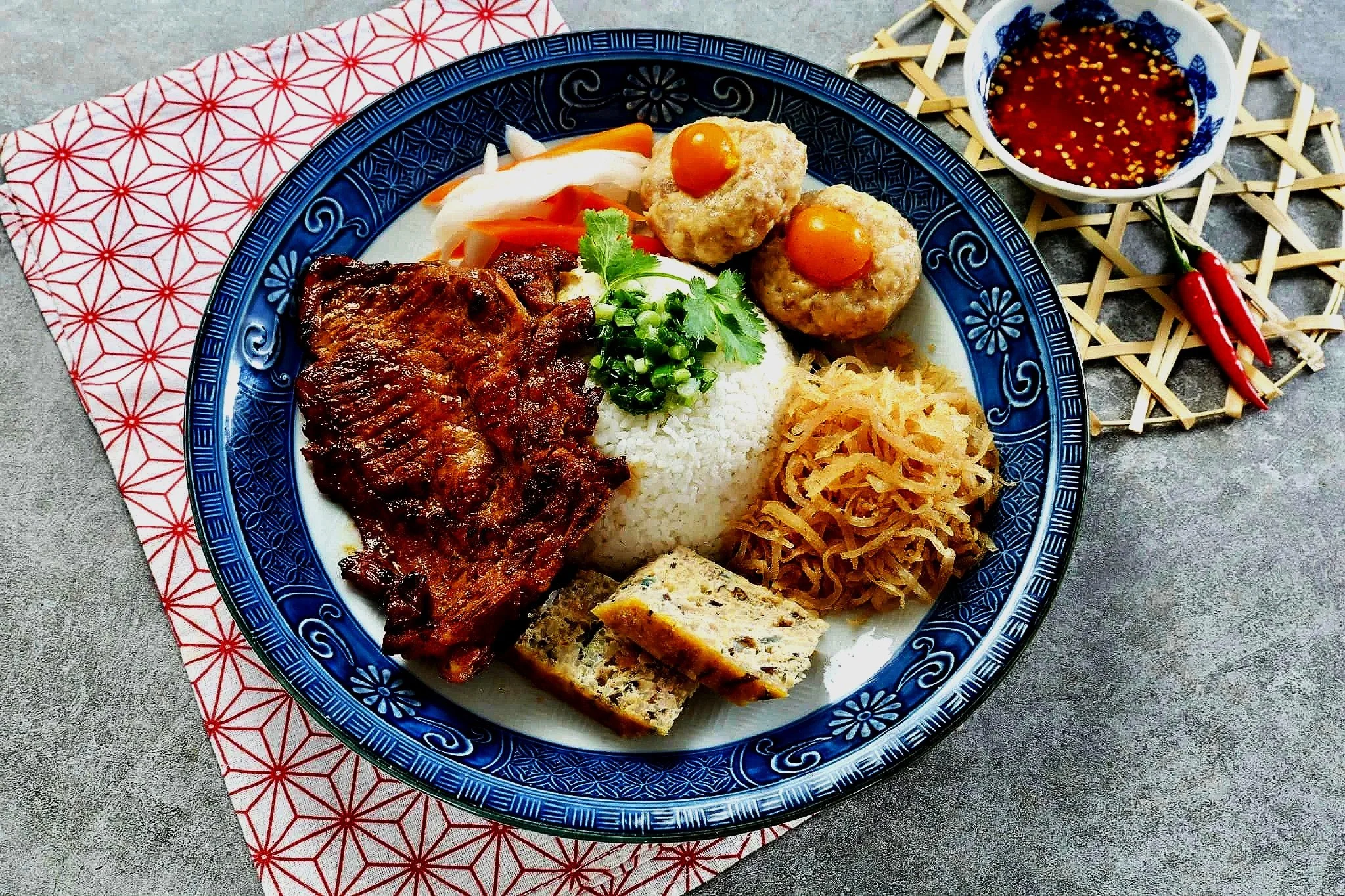 A plate of Asian-style food with rice, grilled meat, pickled vegetables, meatballs with eggs, shredded vegetables, and a slice of tofu, alongside a bowl of dipping sauce and red chili peppers.
