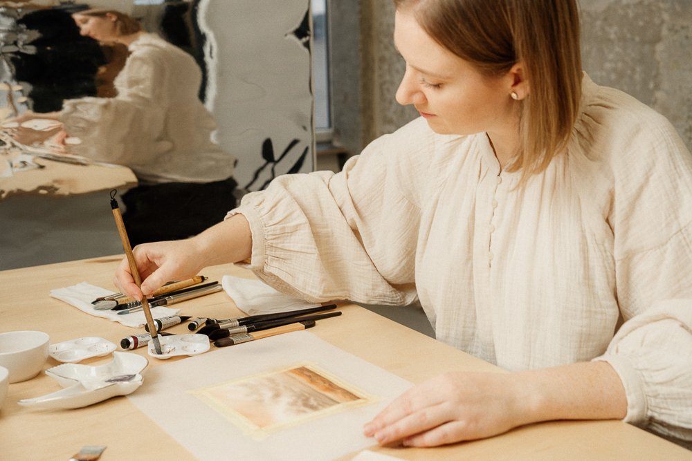 A woman artist painting with brushes and watercolors on a wooden table.