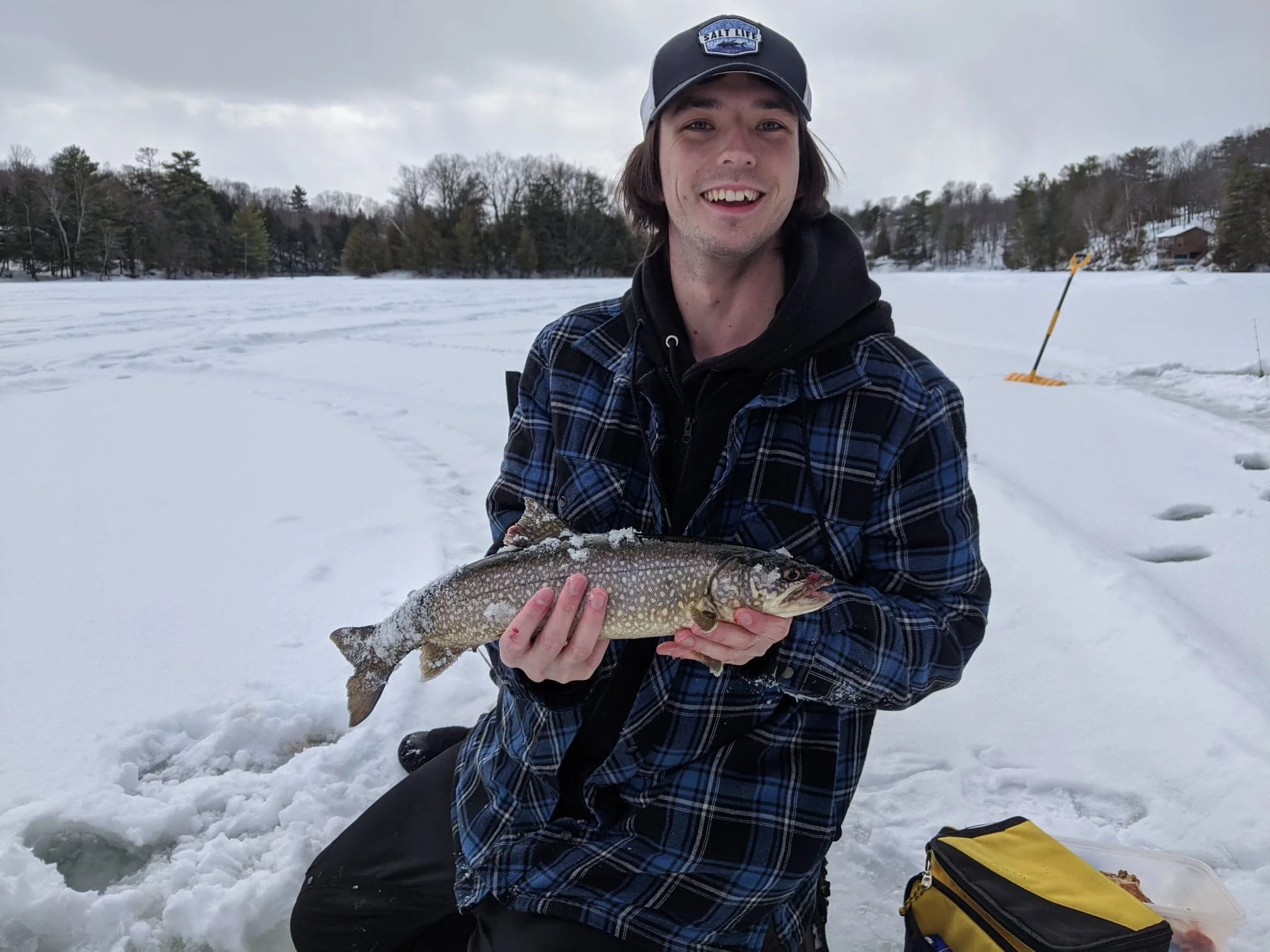 A smiling young man in winter clothing kneels in the snow, holding a freshly caught fish during a winter ice fishing trip on a frozen lake with snow-covered trees in the background.
