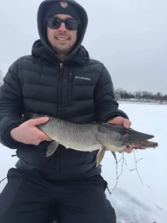 Man wearing a black jacket, sunglasses, and a hat, holding a large fish outdoors in a snowy landscape.
