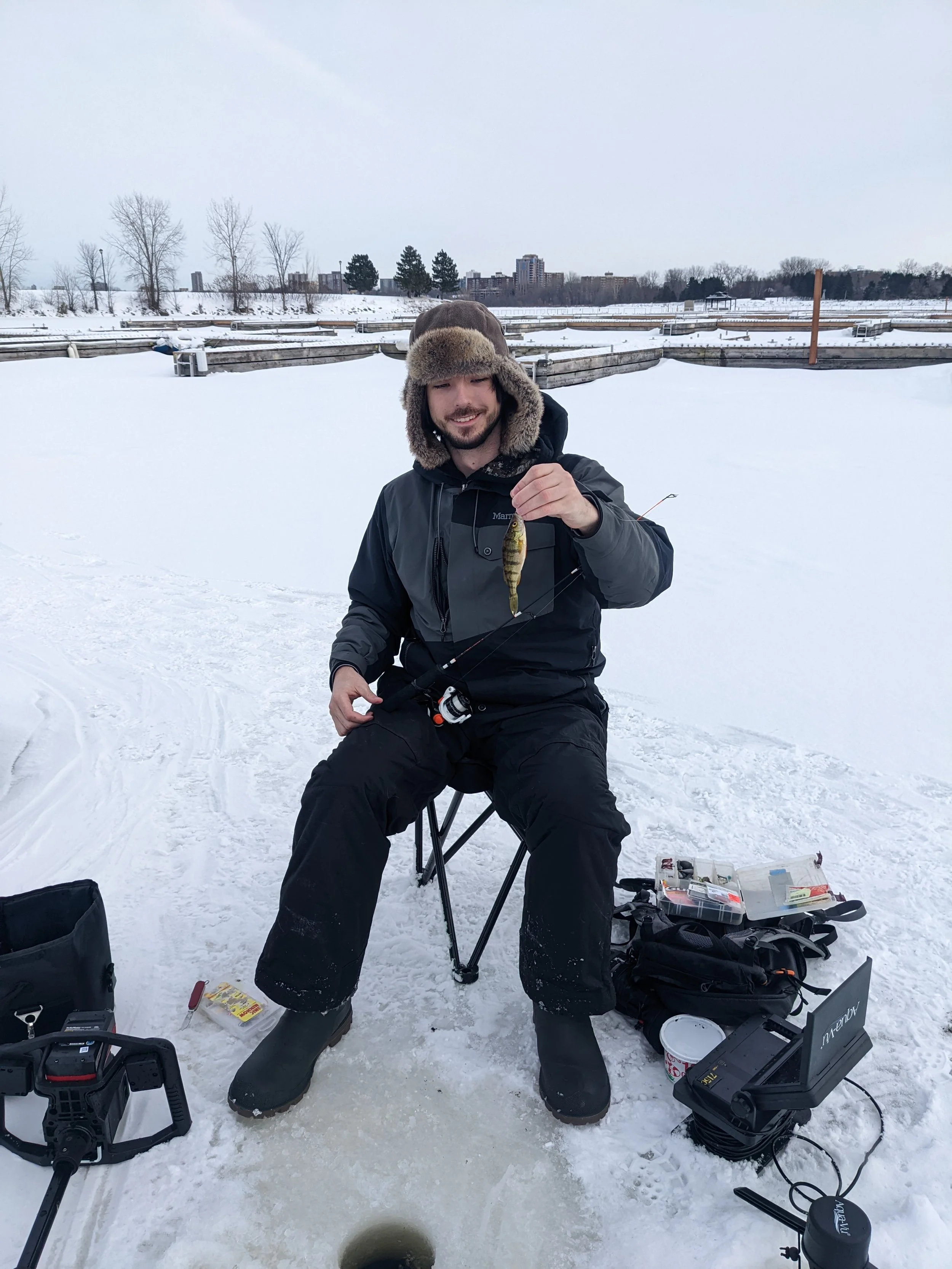 A man sitting on a folding chair on ice during winter, holding a small fish he caught, with fishing gear and supplies around him, snow-covered landscape in the background.
