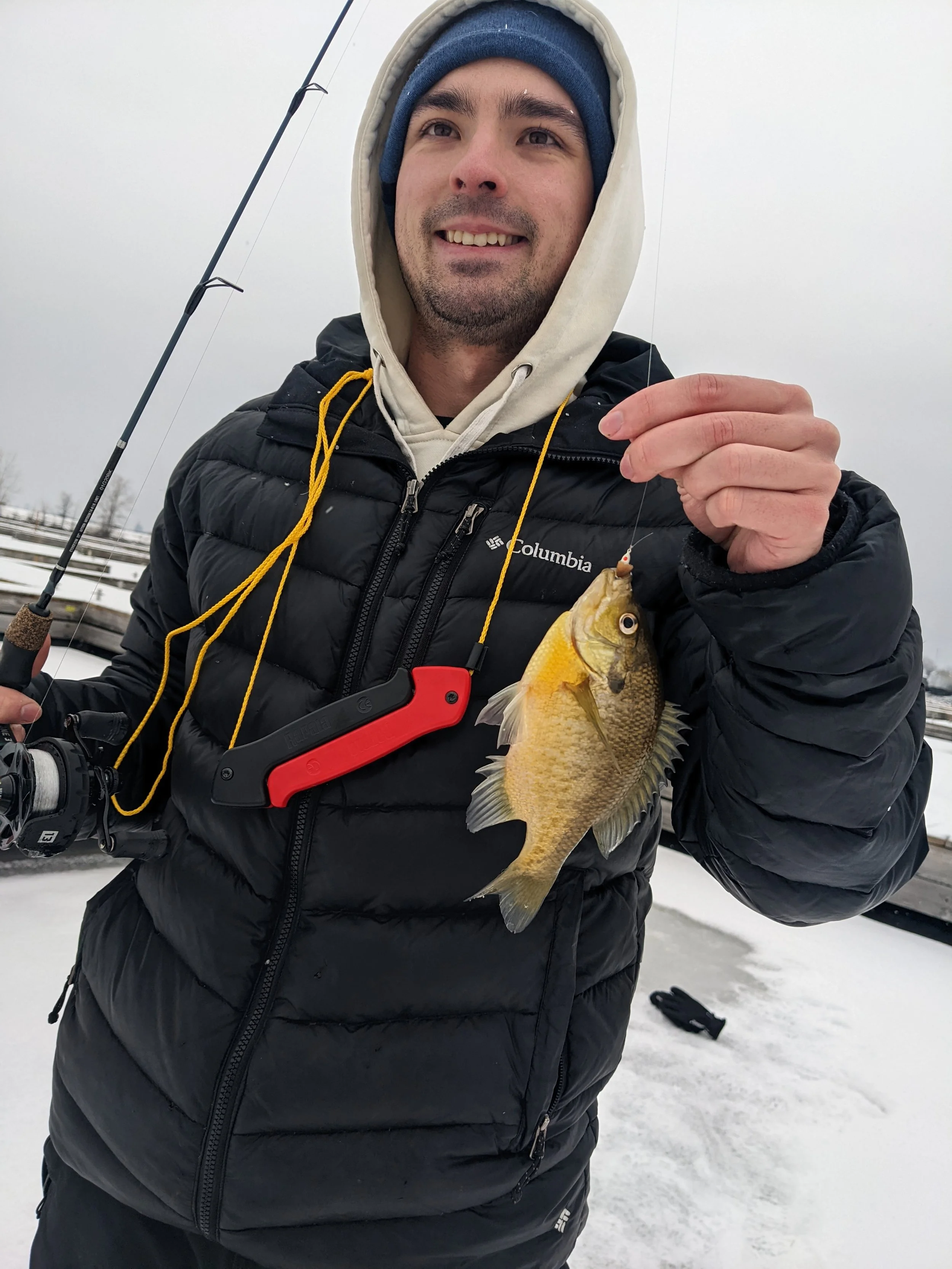 A man dressed in winter clothing holding a fishing rod in one hand and a small fish in the other, standing on snowy ground outdoors.