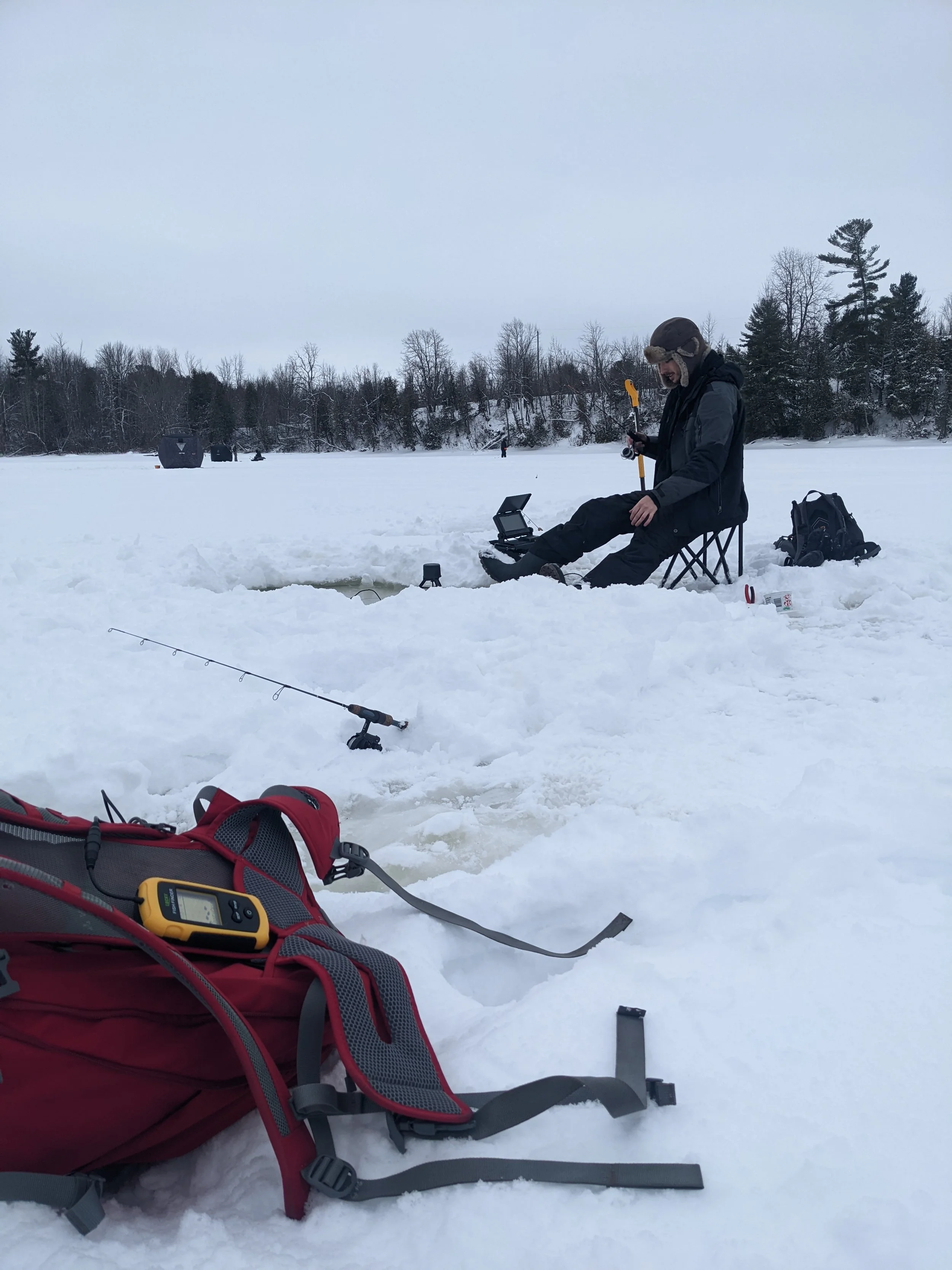 A person ice fishing on a snow-covered lake, sitting in a folding chair with fishing equipment including a rod, auger, and electronic device nearby, with a red backpack on the snow in the foreground.
