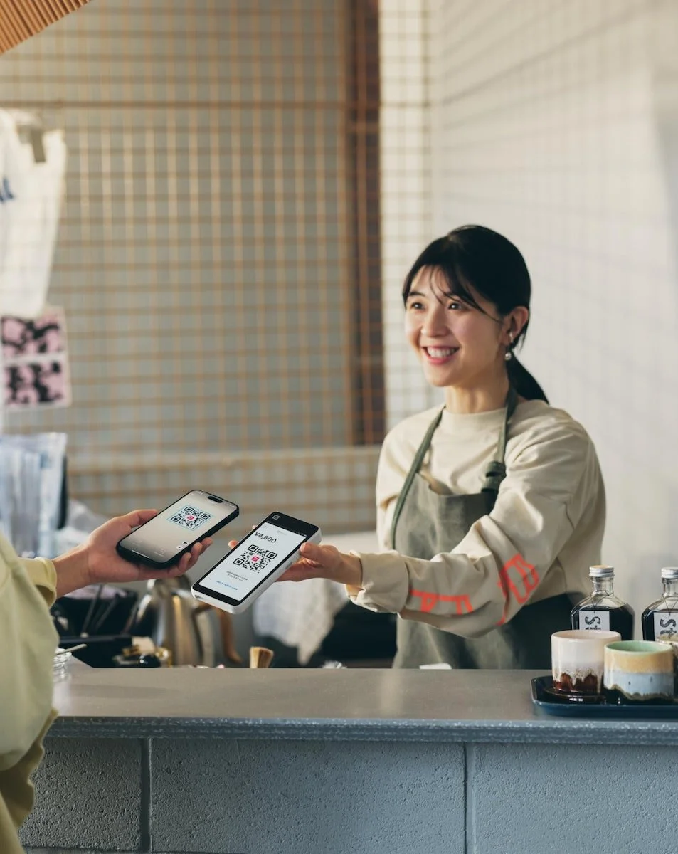 A woman at a coffee shop counter, smiling and handing a smartphone with a QR code to a customer, with another smartphone showing the same QR code. There are bottles and cups on the counter.