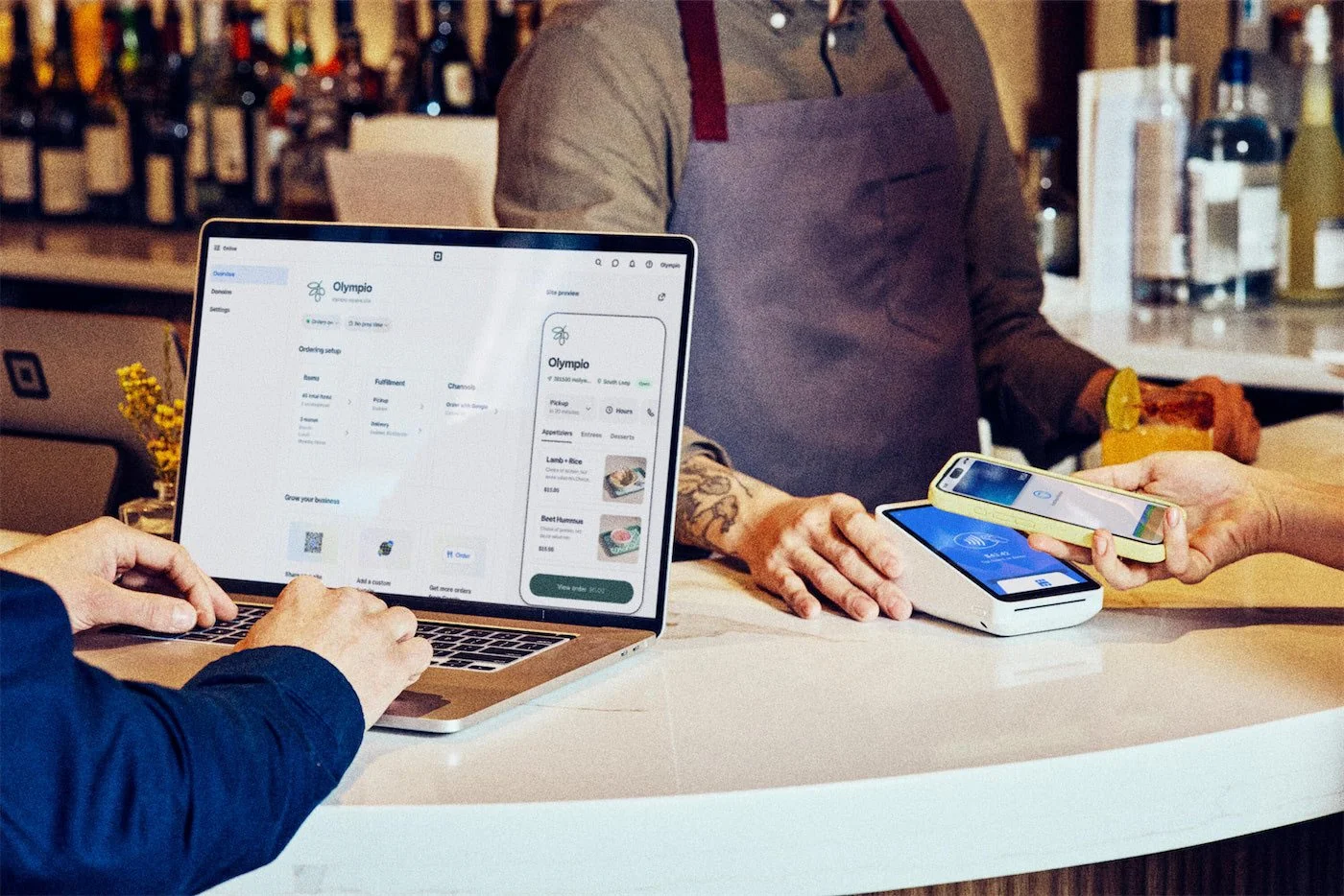 Person using a laptop at a counter, with a second person holding a mobile phone with contactless payment, and a third person standing nearby with a drink.