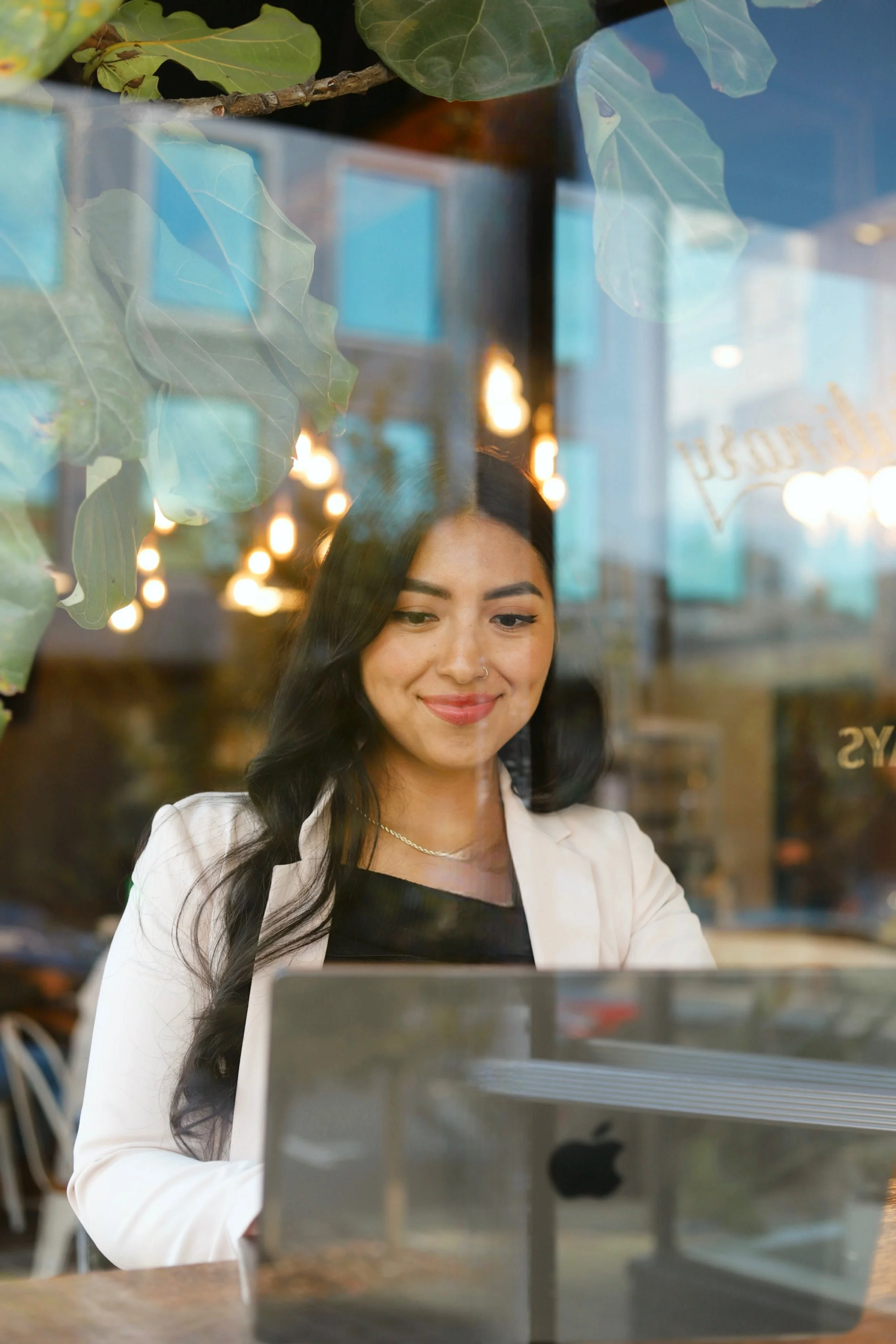 A woman with long dark hair, wearing a white blazer and black top, smiling while working on a laptop in a cozy café. The photo is taken through a window with reflections of interior lights and some green leaves.
