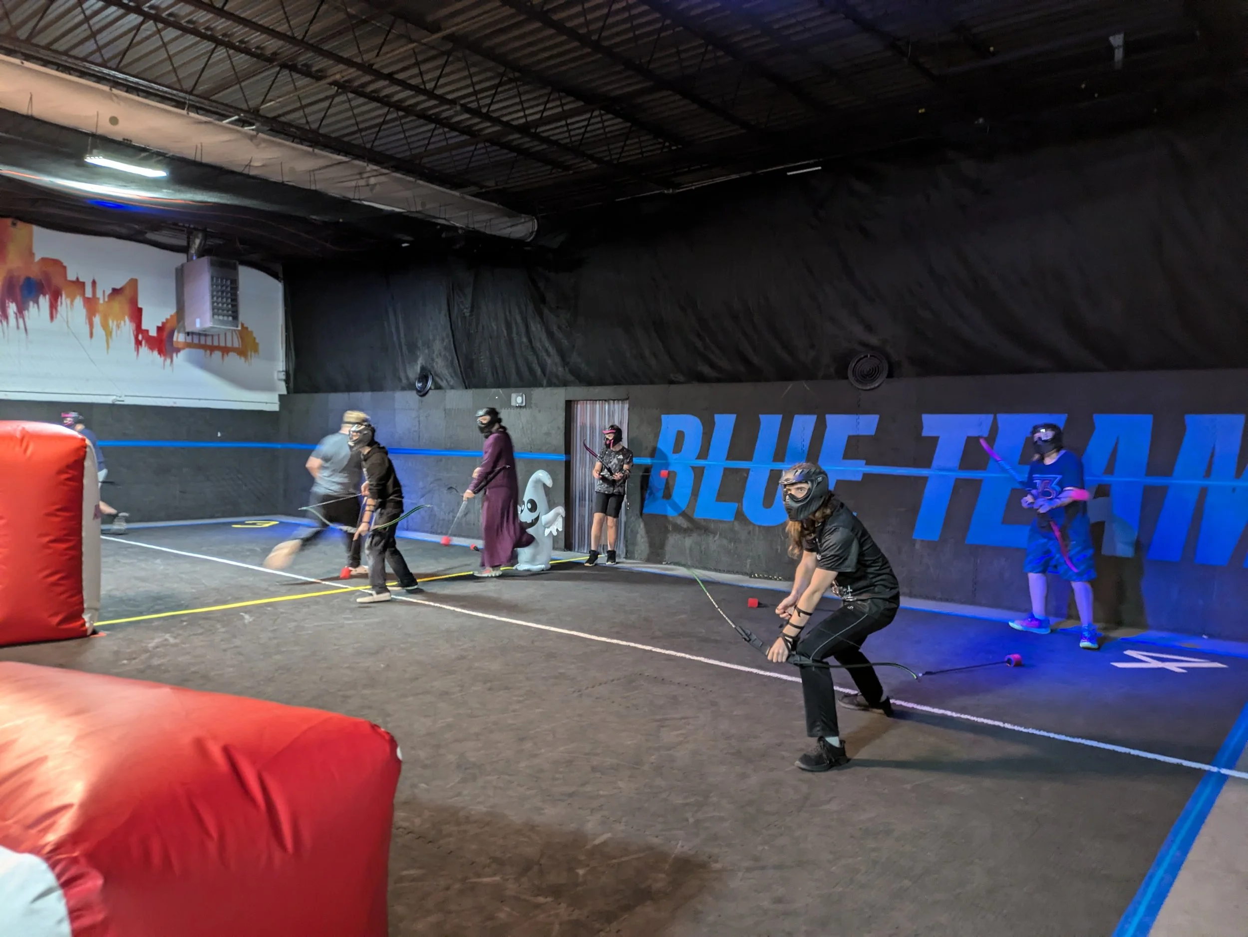 People wearing protective gear playing laser tag in an indoor arena with a black wall that reads 'BLUE TEAM' and a cityscape mural in the background.