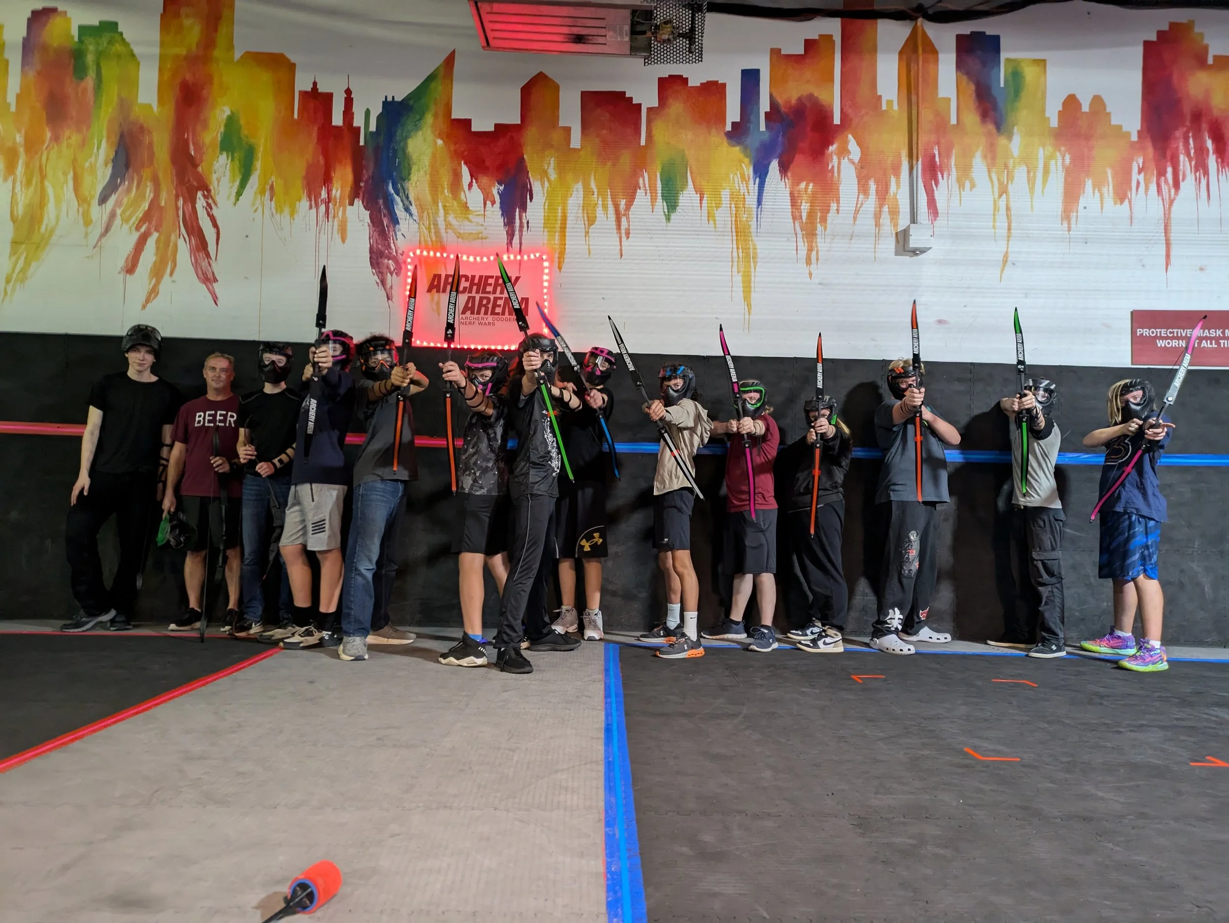 A group of children and teenagers at an indoor archery range, holding bows and arrows, standing in front of a colorful mural of a city skyline painted on the wall, with two adult supervisors on each side. The floor is marked with colored lines for shooting lanes.