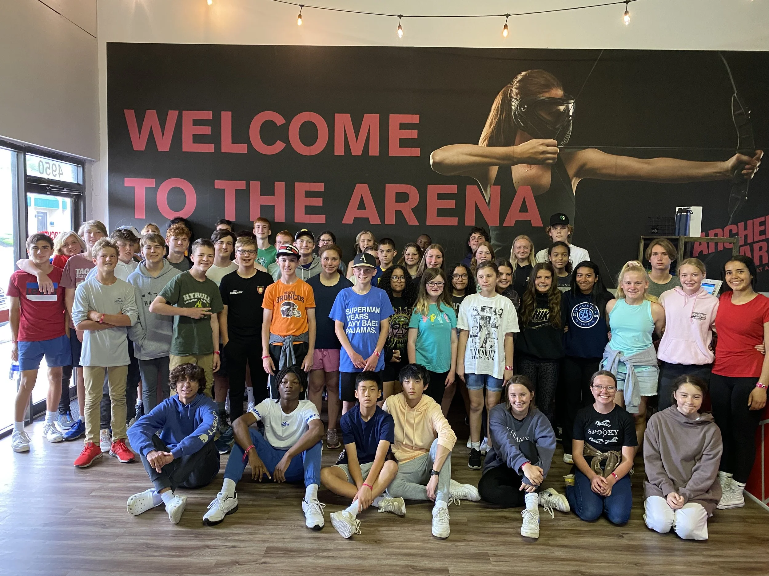 Group of students posing in front of a large wall with a mural of a woman drawing a bow and the words "Welcome to the Arena" during daytime.