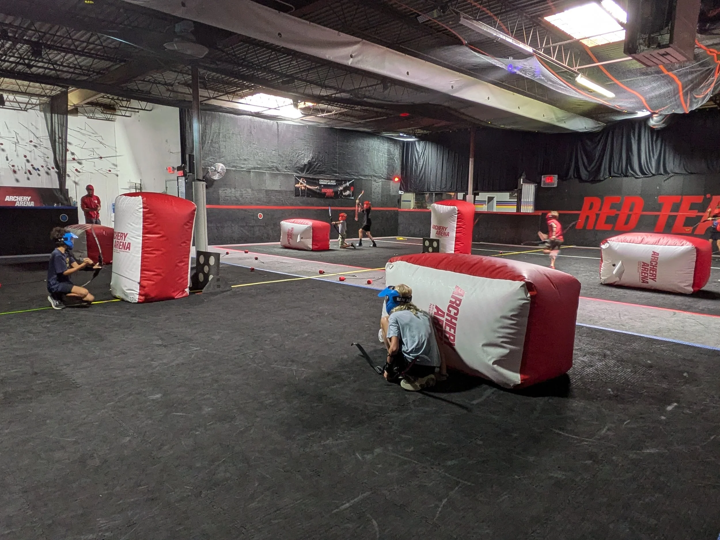 Indoor dodgeball arena with players, padded walls, and inflatable barriers, with people wearing protective helmets and using rackets.