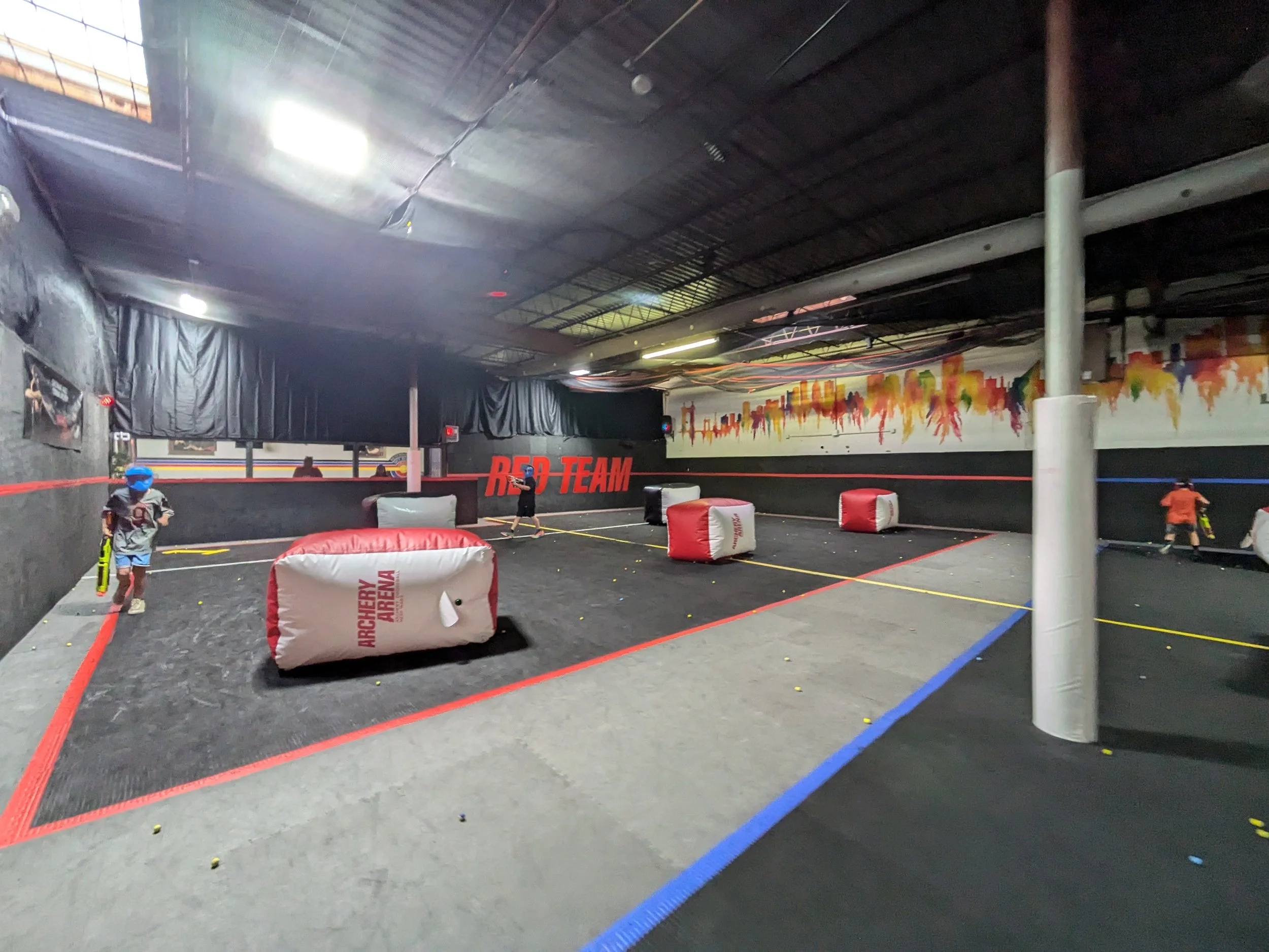 Indoor archery arena with padded barriers, yellow and blue boundary lines, graffiti-style wall art, and children practicing archery.