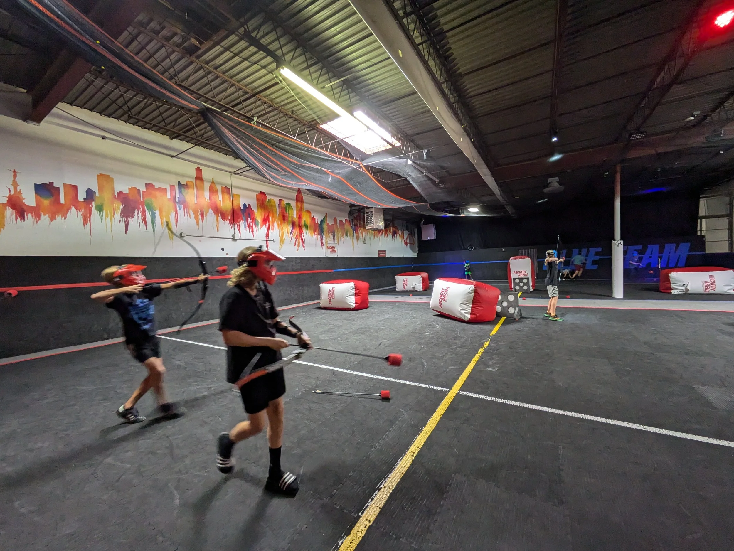 Indoor laser tag or archery tag arena with children wearing red helmets and protective gear, aiming bows and arrows. The arena has a black floor, colorful wall art, inflatable barriers, and neon lighting.