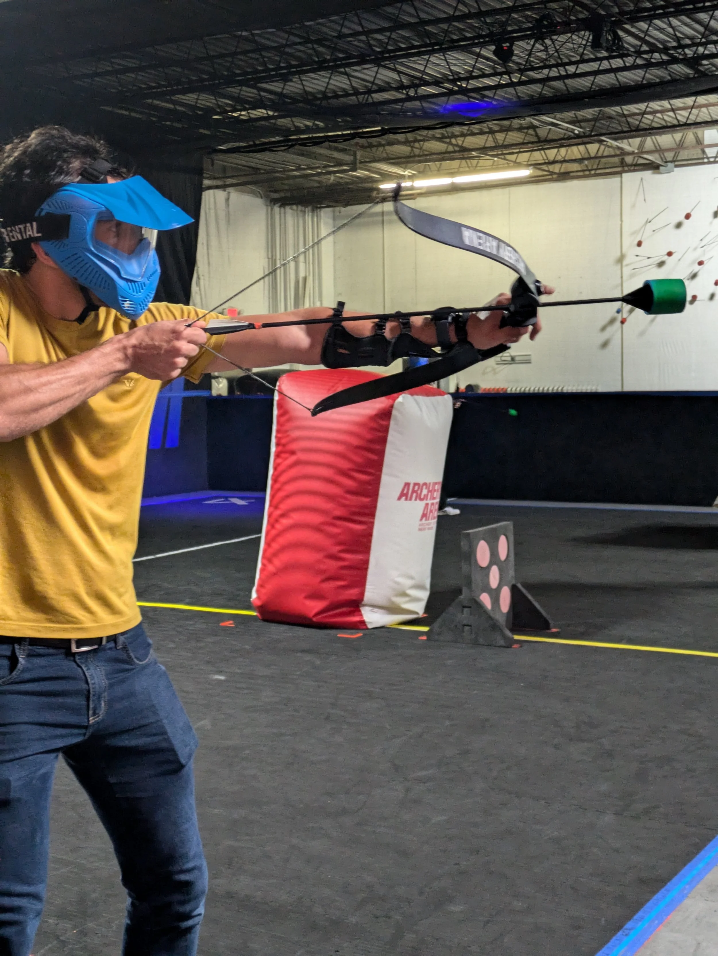 A man wearing a blue face shield, yellow shirt, and jeans practicing archery indoors, aiming with a black bow at targets.