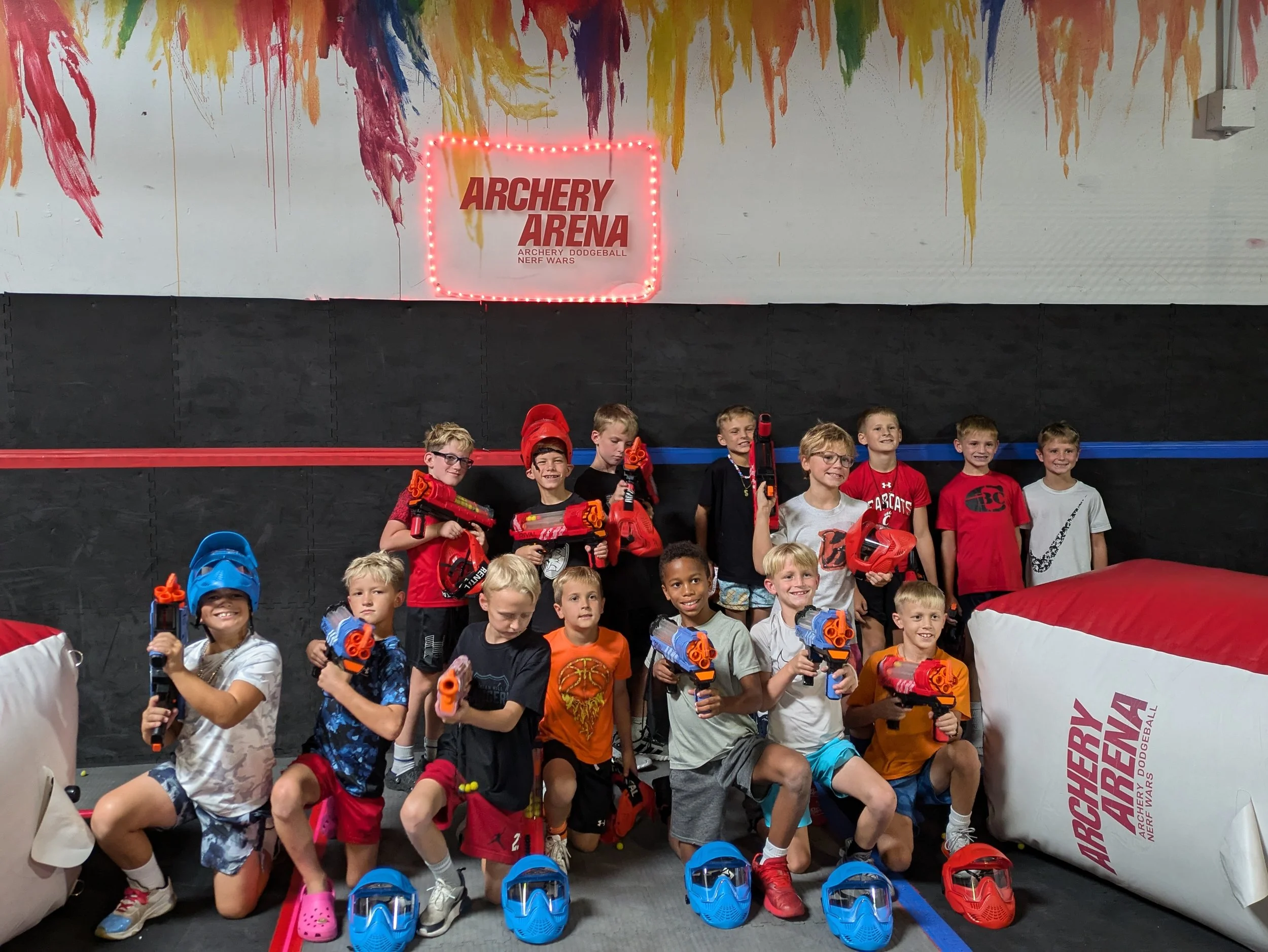 Group of kids at indoor laser tag arena holding toy guns, wearing protective gear, and smiling.