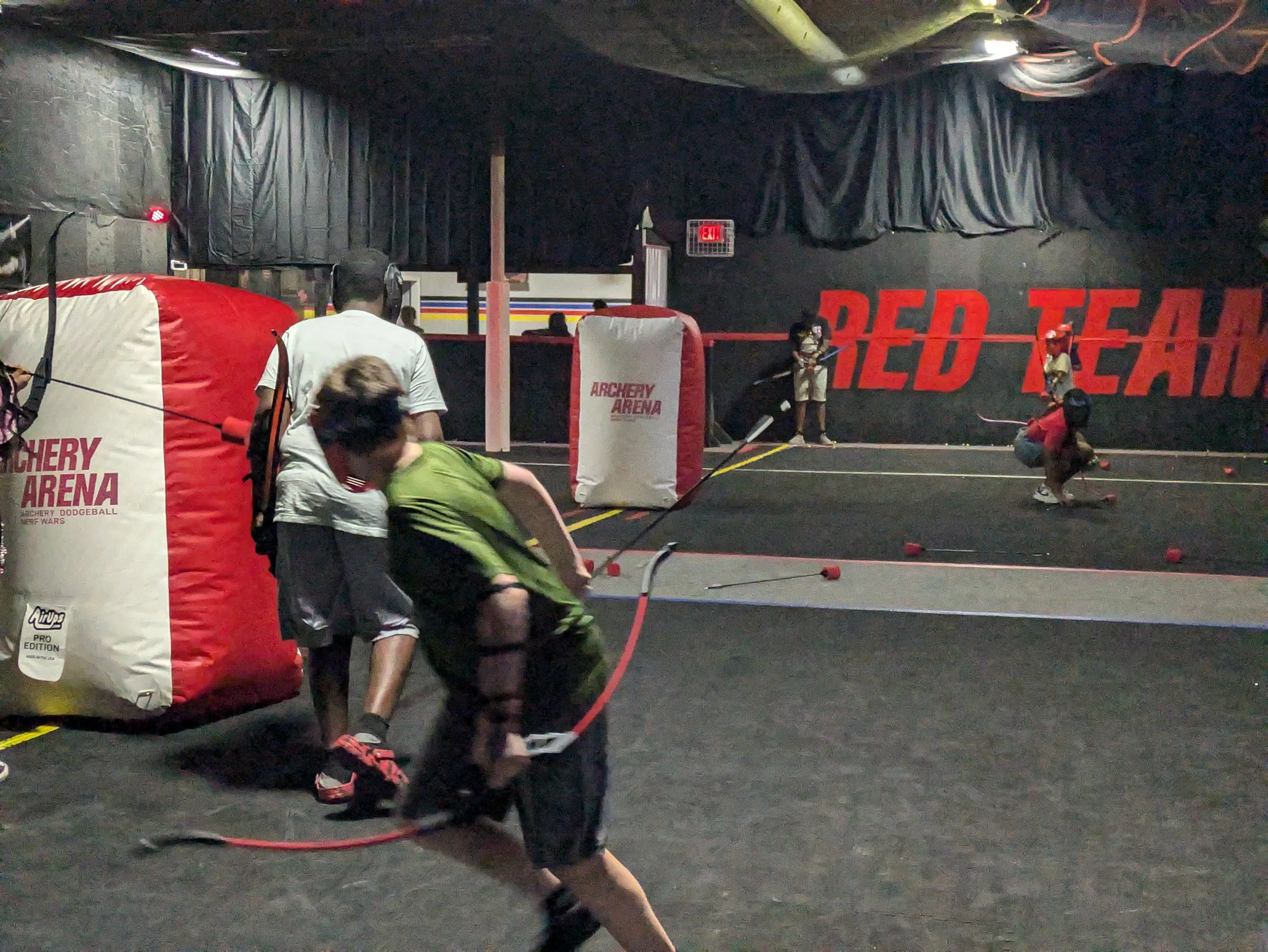 Children practicing archery at an indoor archery arena, with a black wall displaying 'RED TEAM' in large red letters in the background.