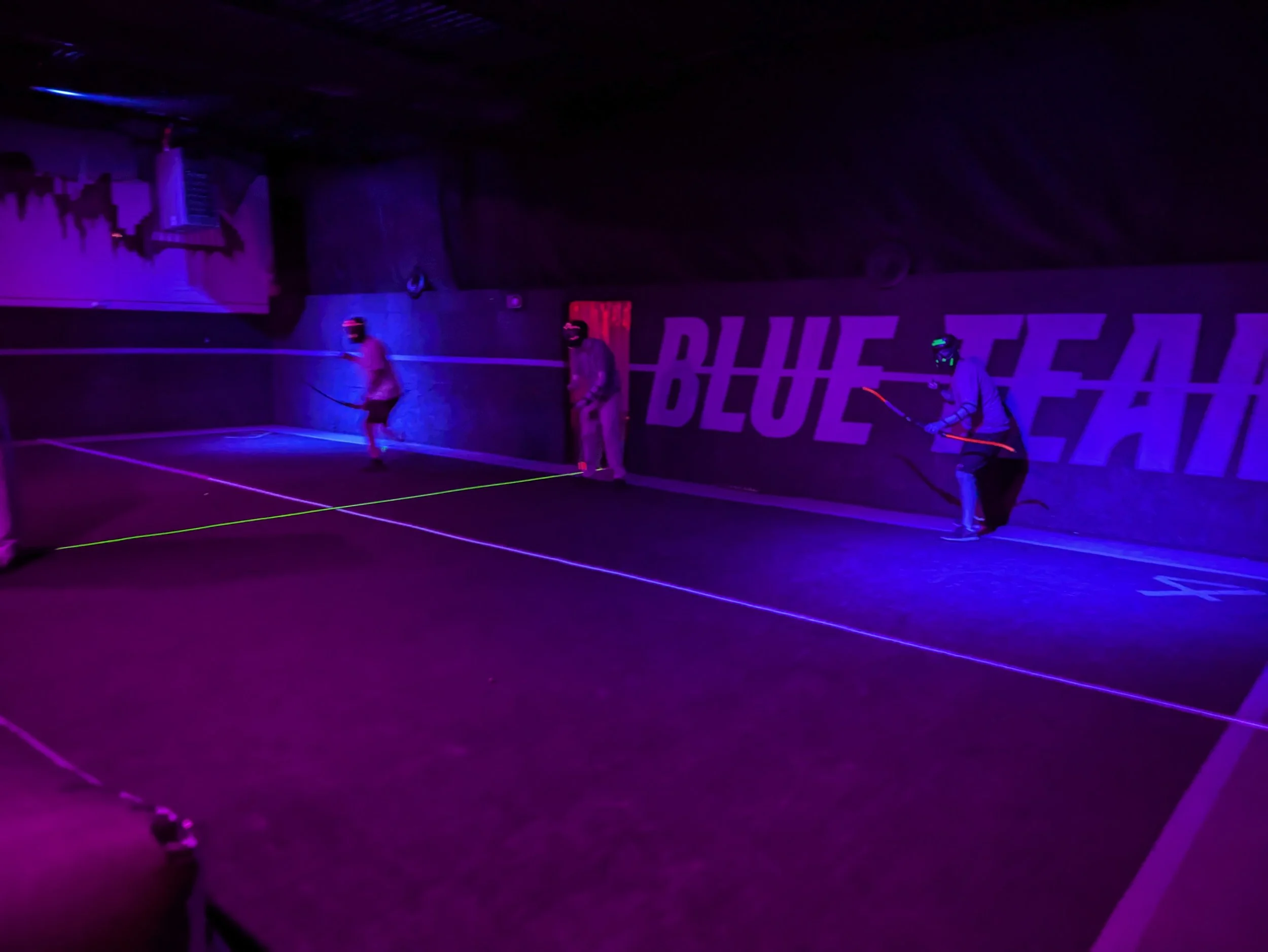 Three people playing glow-in-the-dark archery. The room is dimly lit with blue and purple lighting, and the players are wearing protective gear with glowing accents.