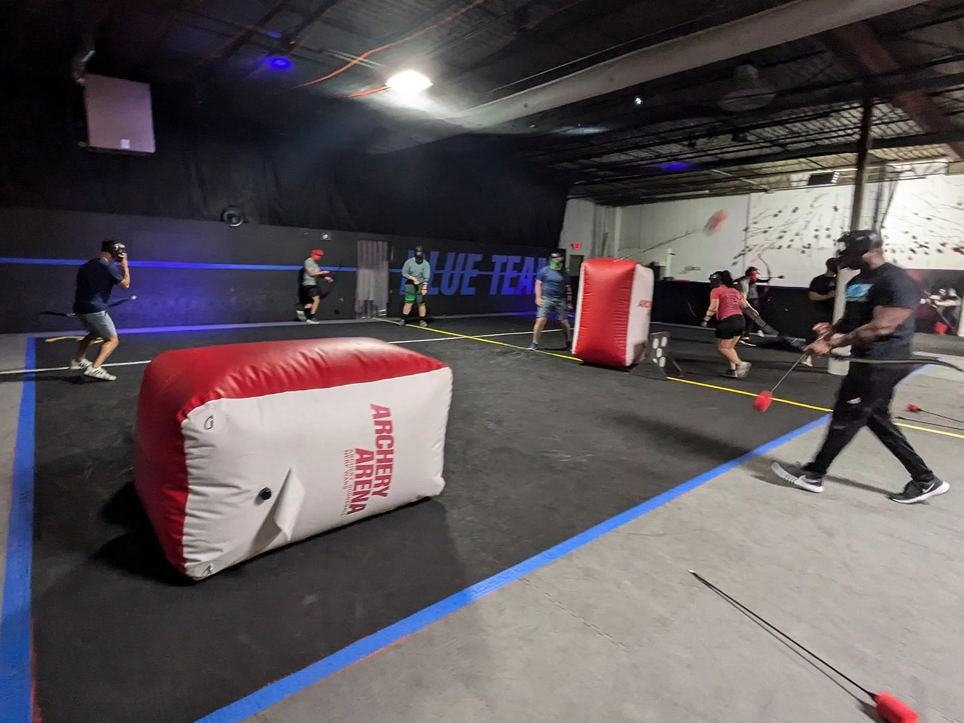 People playing archery tag in an indoor facility, aiming at targets with large padded barriers and a climbing wall in the background.